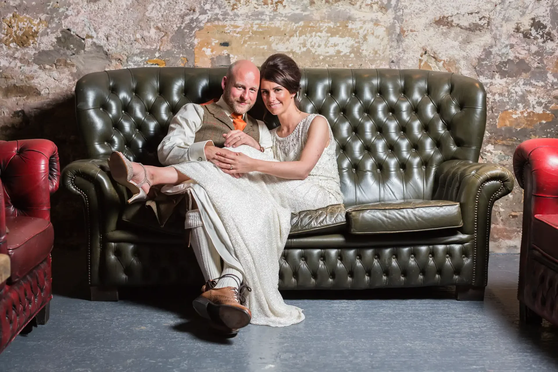 A couple in formal attire sitting closely on a leather sofa, smiling, in a room with rustic walls.