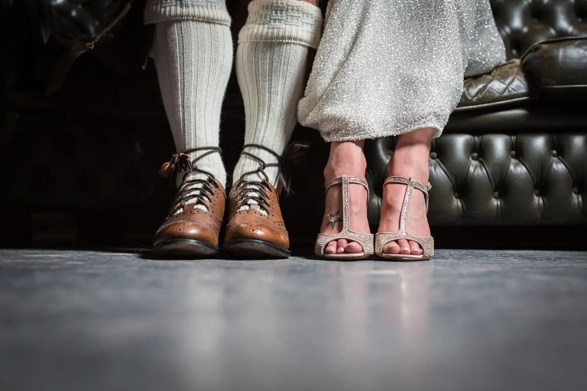 Two people standing side by side showcasing their footwear, one in brown lace-up shoes with socks, the other in silver strappy sandals, against a dark floor background.