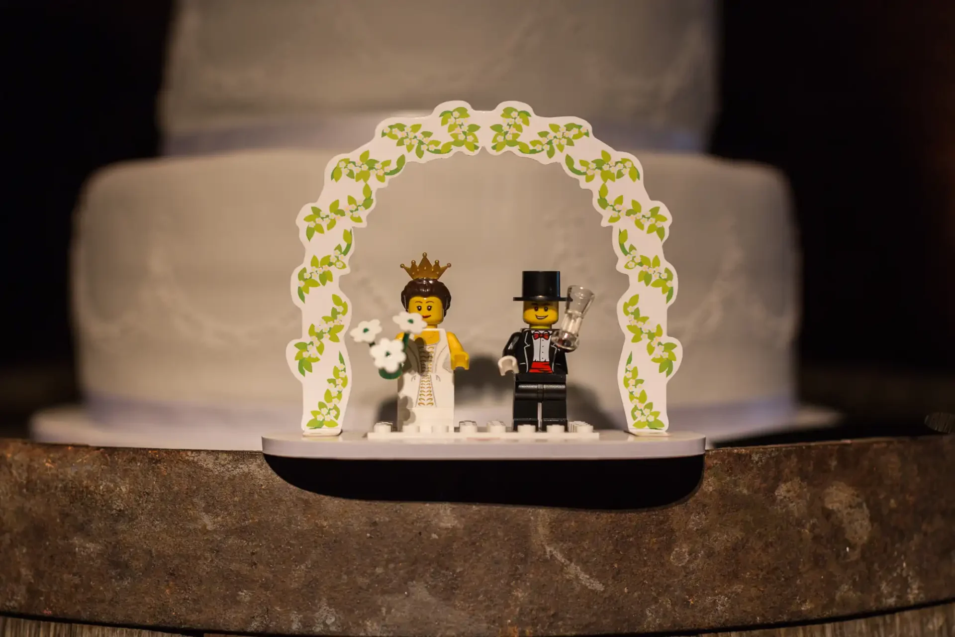 Lego bride and groom figures under a floral arch, with a blurred wedding cake in the background.