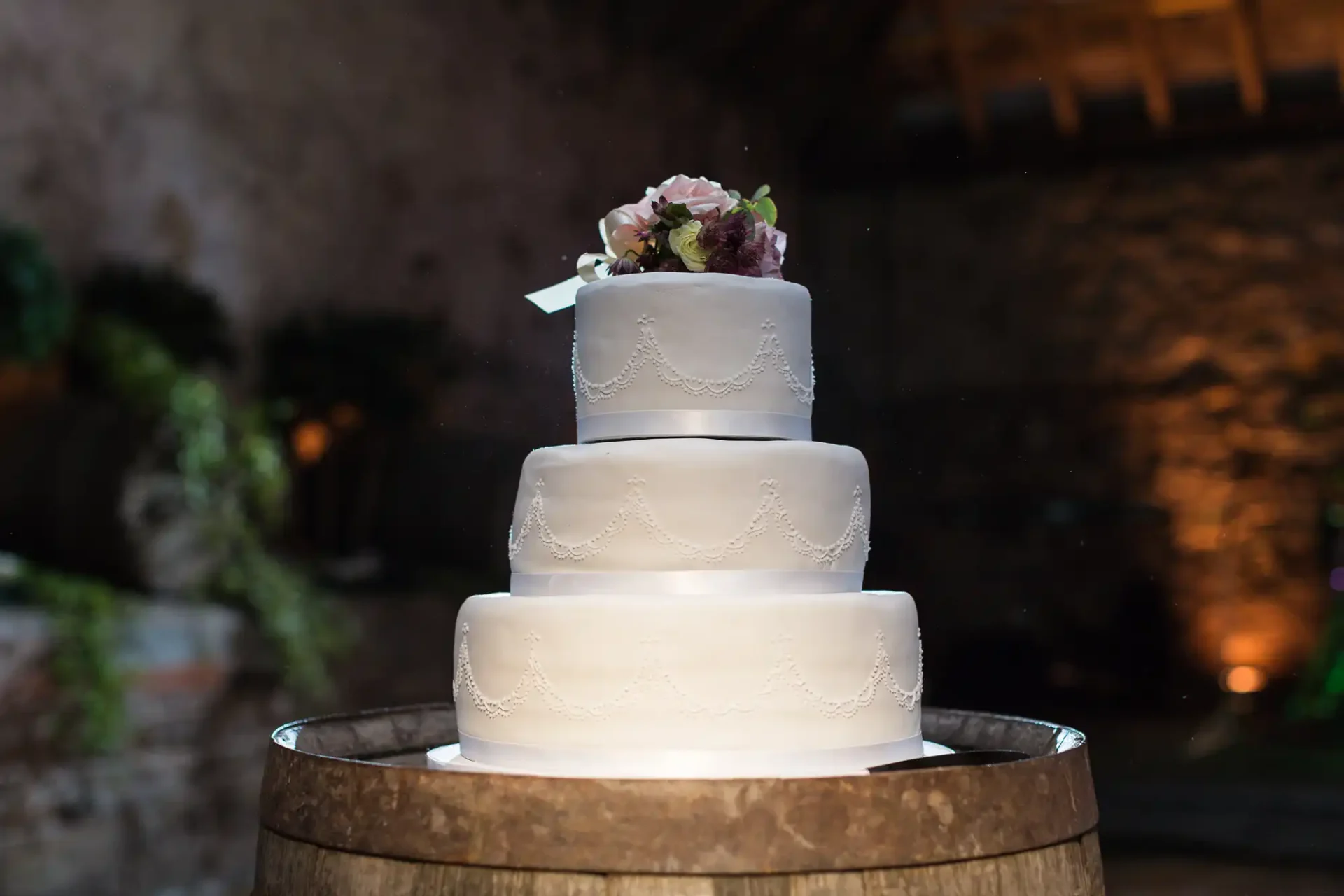 Three-tiered wedding cake with white icing and floral decoration on top, displayed on a wooden barrel in a dimly lit setting.
