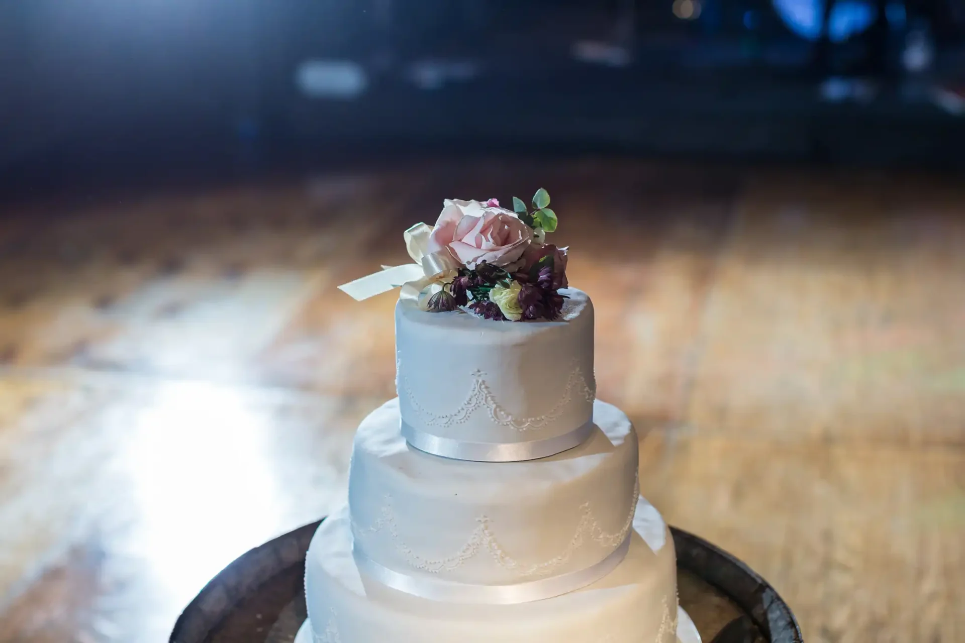 A three-tiered wedding cake with white frosting and floral decorations on top, displayed on a wooden table with a softly lit background.