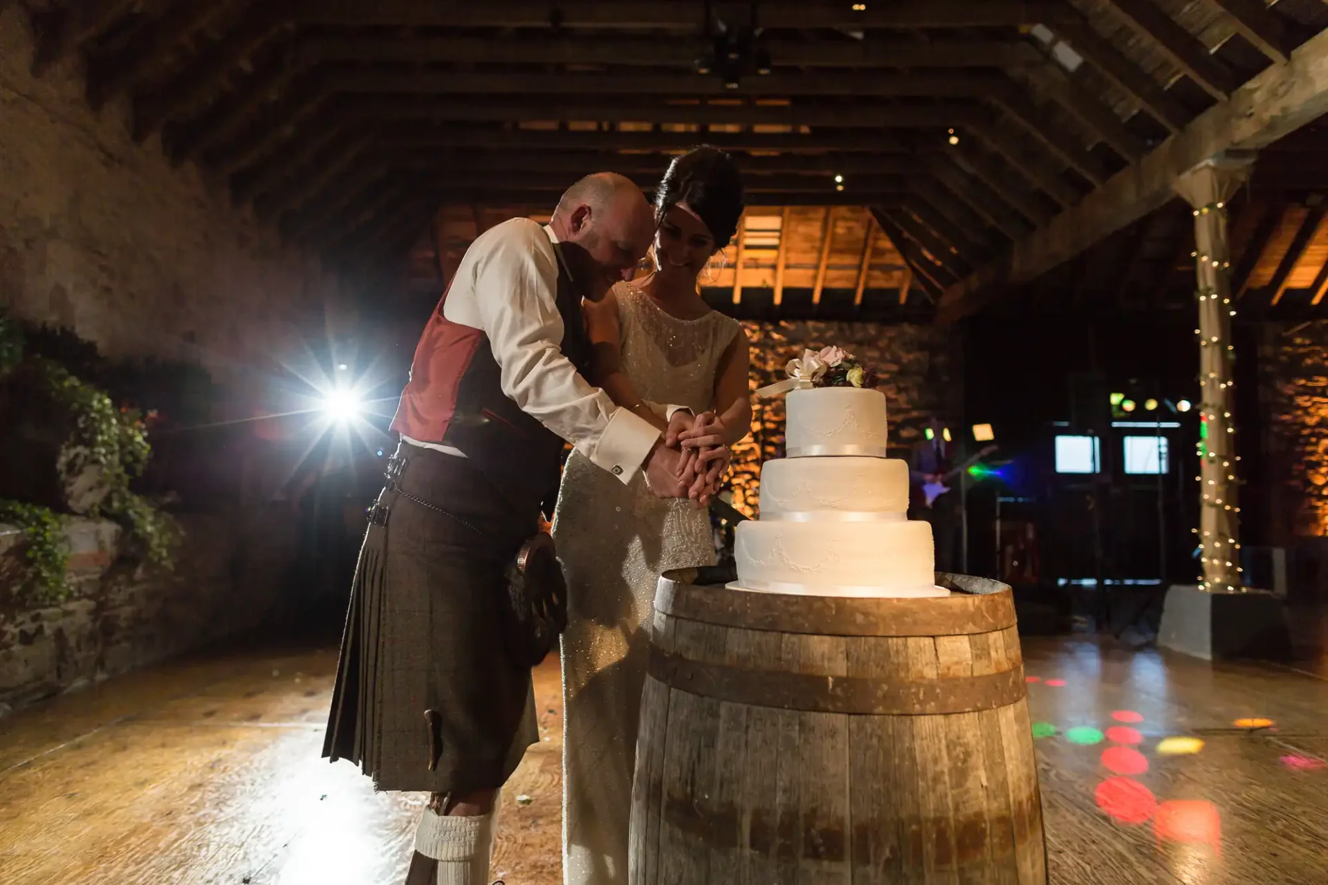 A bride and groom in formal attire cut a wedding cake together in a rustic barn venue, with warm lighting and a lens flare.