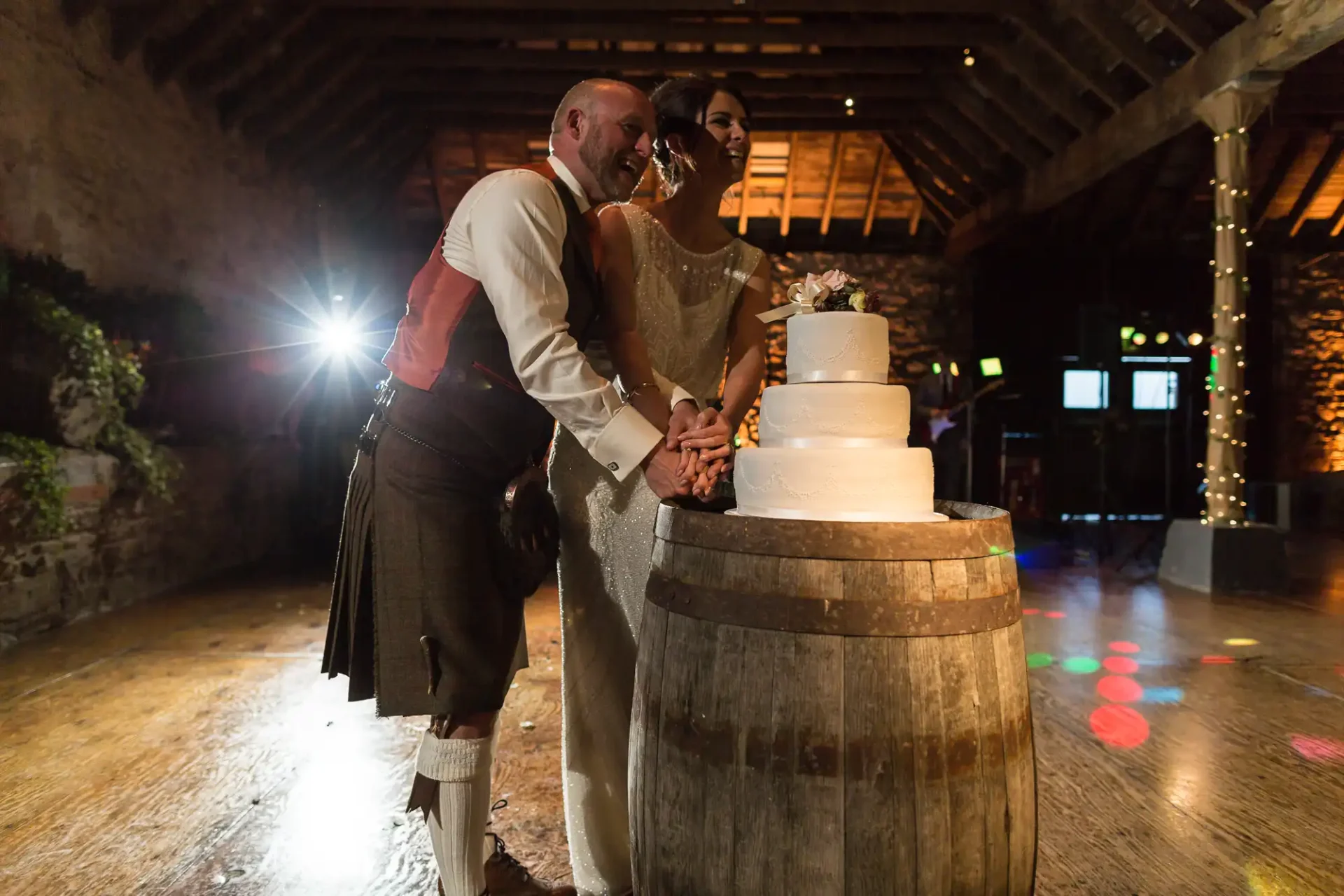 A couple in wedding attire, the man in a kilt, cutting a cake on a barrel in a dimly lit rustic room with festive lights.