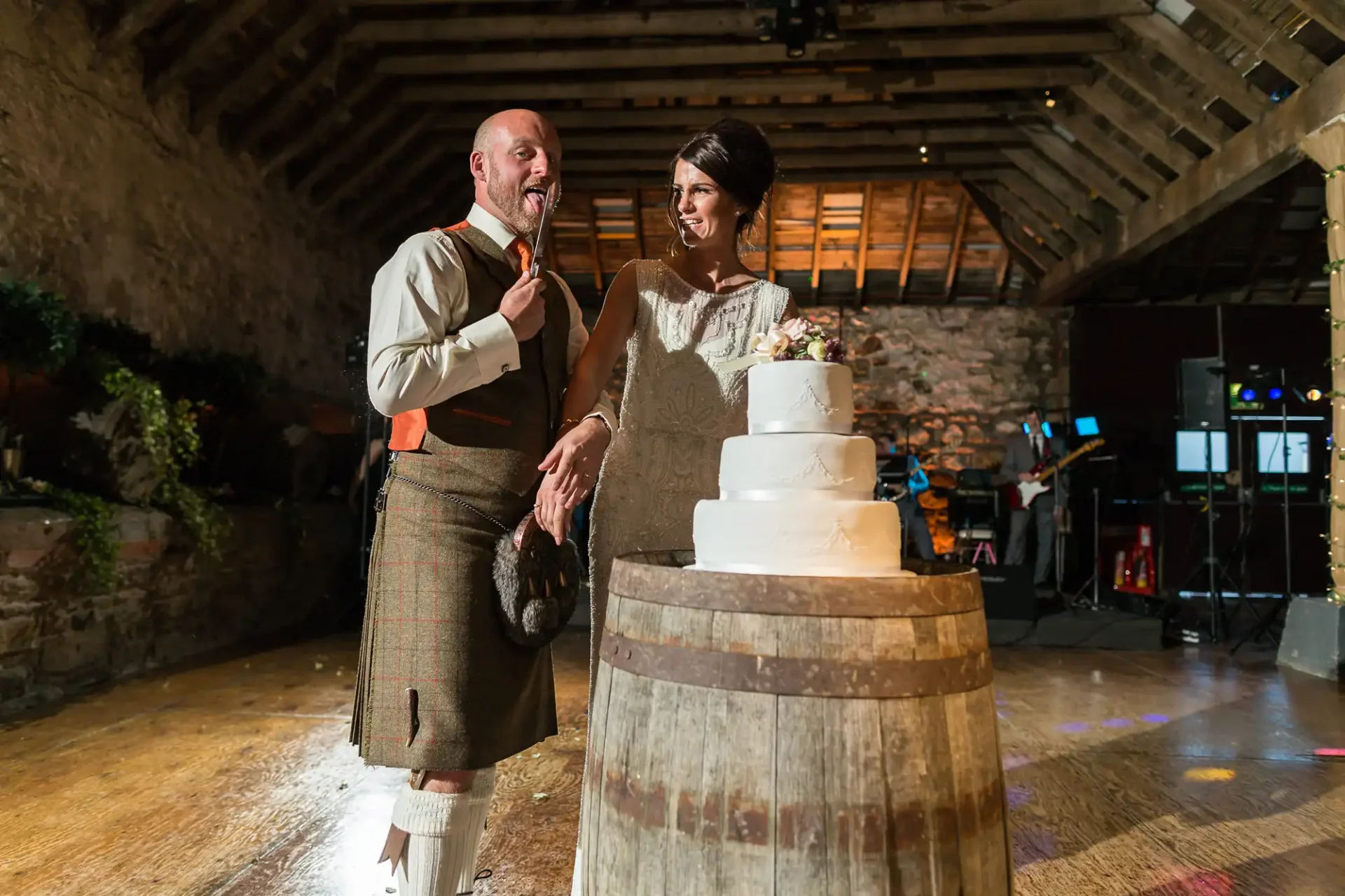 A bride and groom in a rustic venue with the groom in a kilt, laughing while holding a knife over a wedding cake on a barrel.