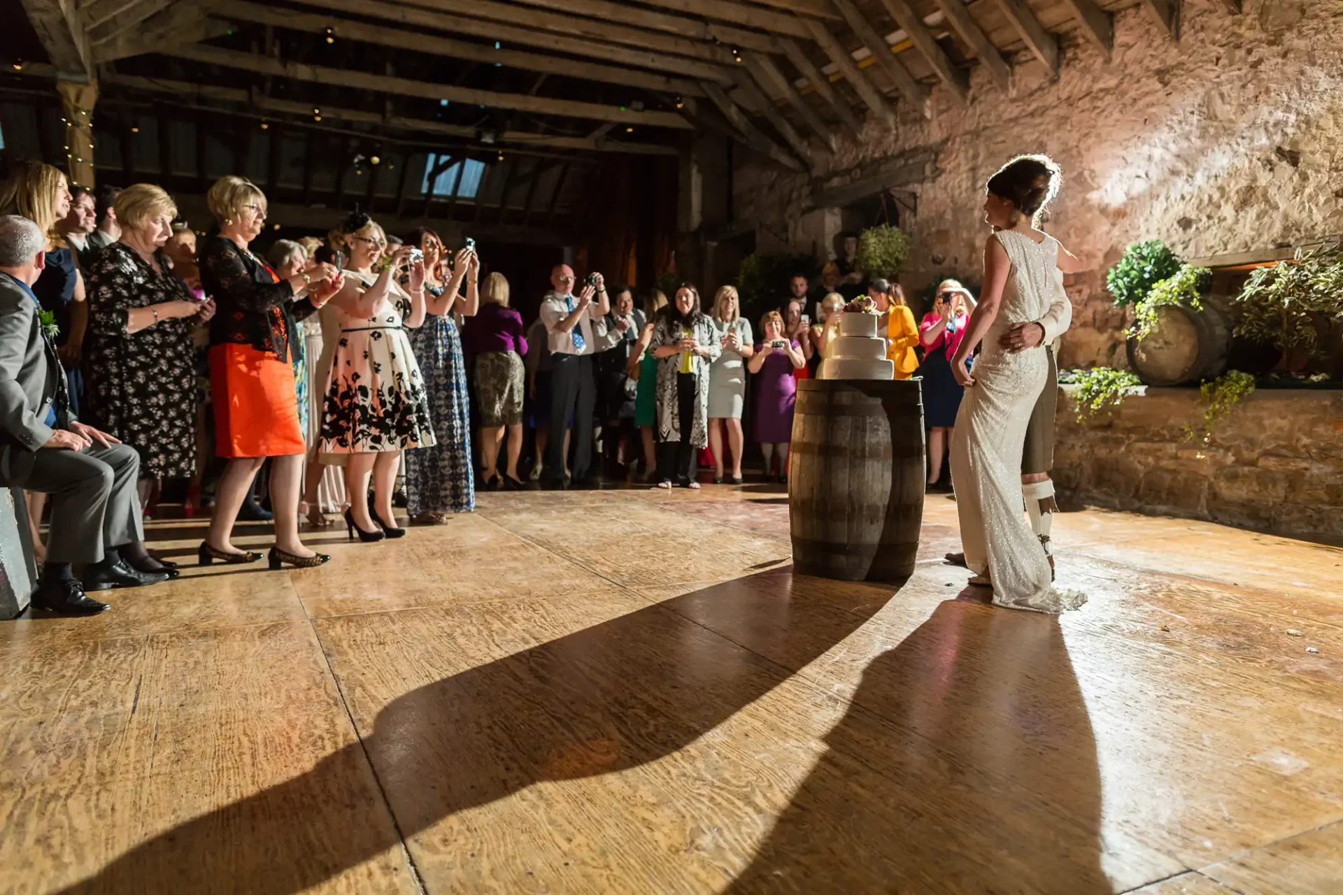A happy bride and groom standing next to a colorful cake, surrounded by applauding guests in a warmly-lit barn.