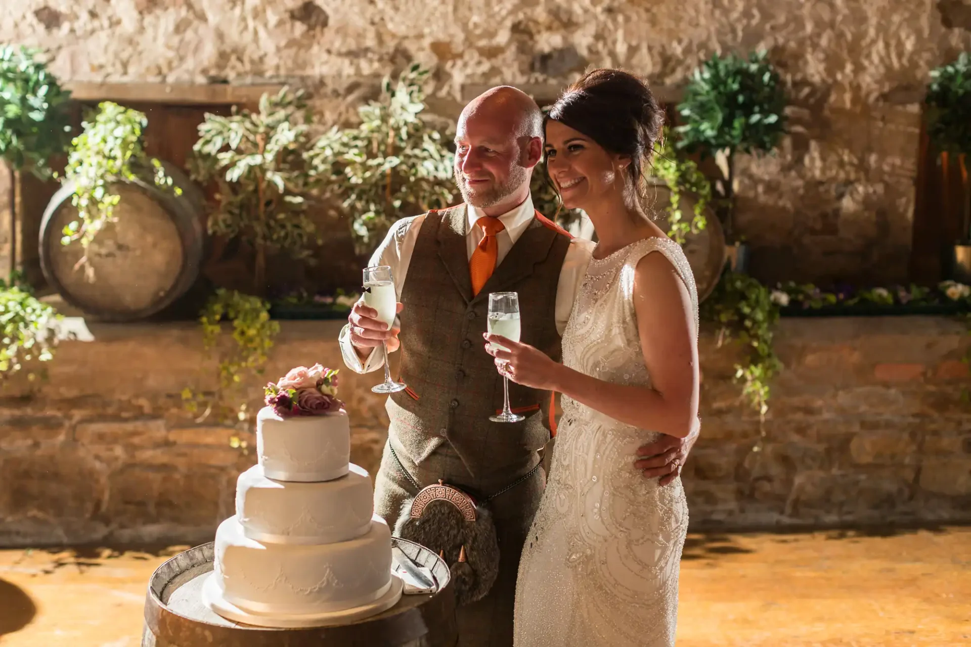 A bride and groom smiling and holding champagne flutes next to a wedding cake in a rustic indoor setting.