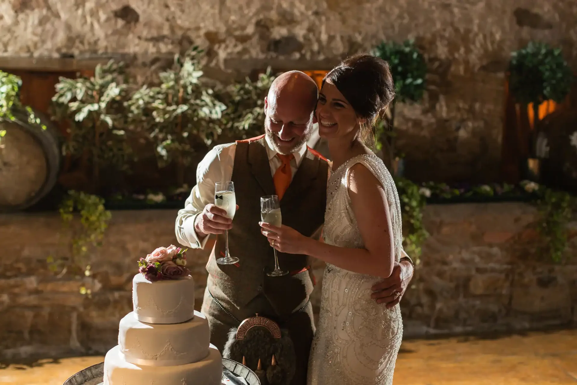 A bride and groom smiling and toasting with champagne glasses beside a wedding cake in a rustic venue lit by warm lights.