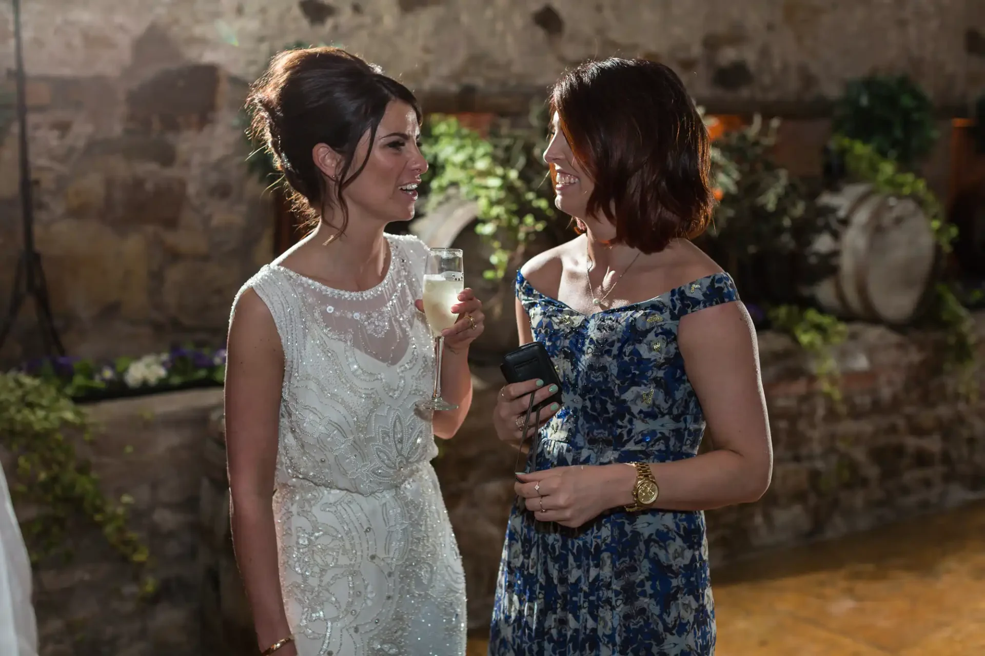Two women smiling and chatting at a formal event, one holding a champagne glass and the other holding a camera.