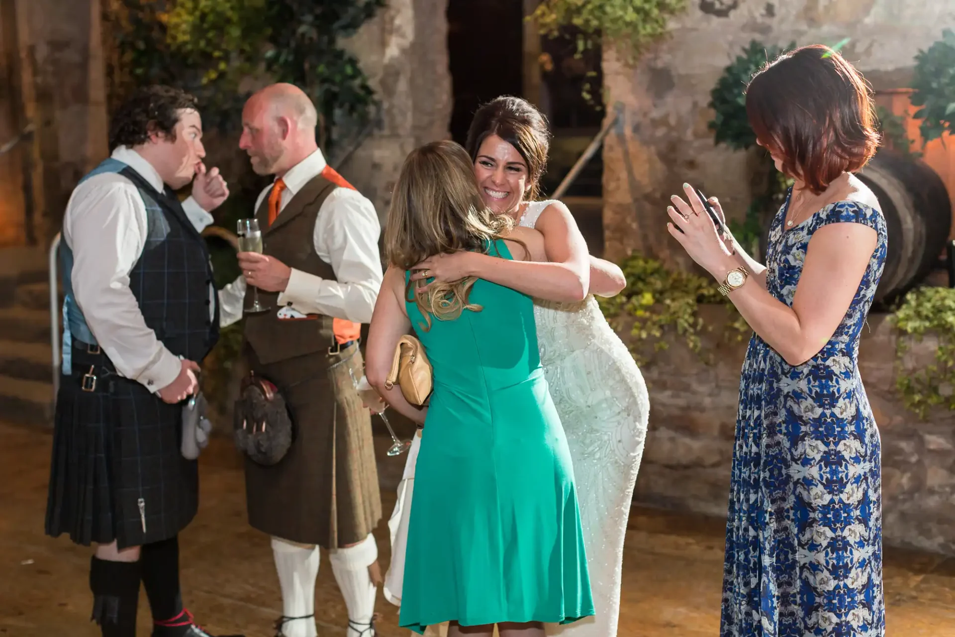 A joyful bride in a white dress hugs a woman in a green dress at an evening reception with three other guests smiling and conversing in the background.