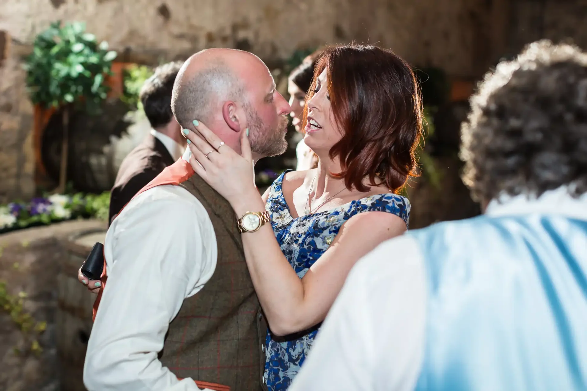 A couple kisses affectionately at an event, surrounded by guests in a rustic, plant-decorated setting.