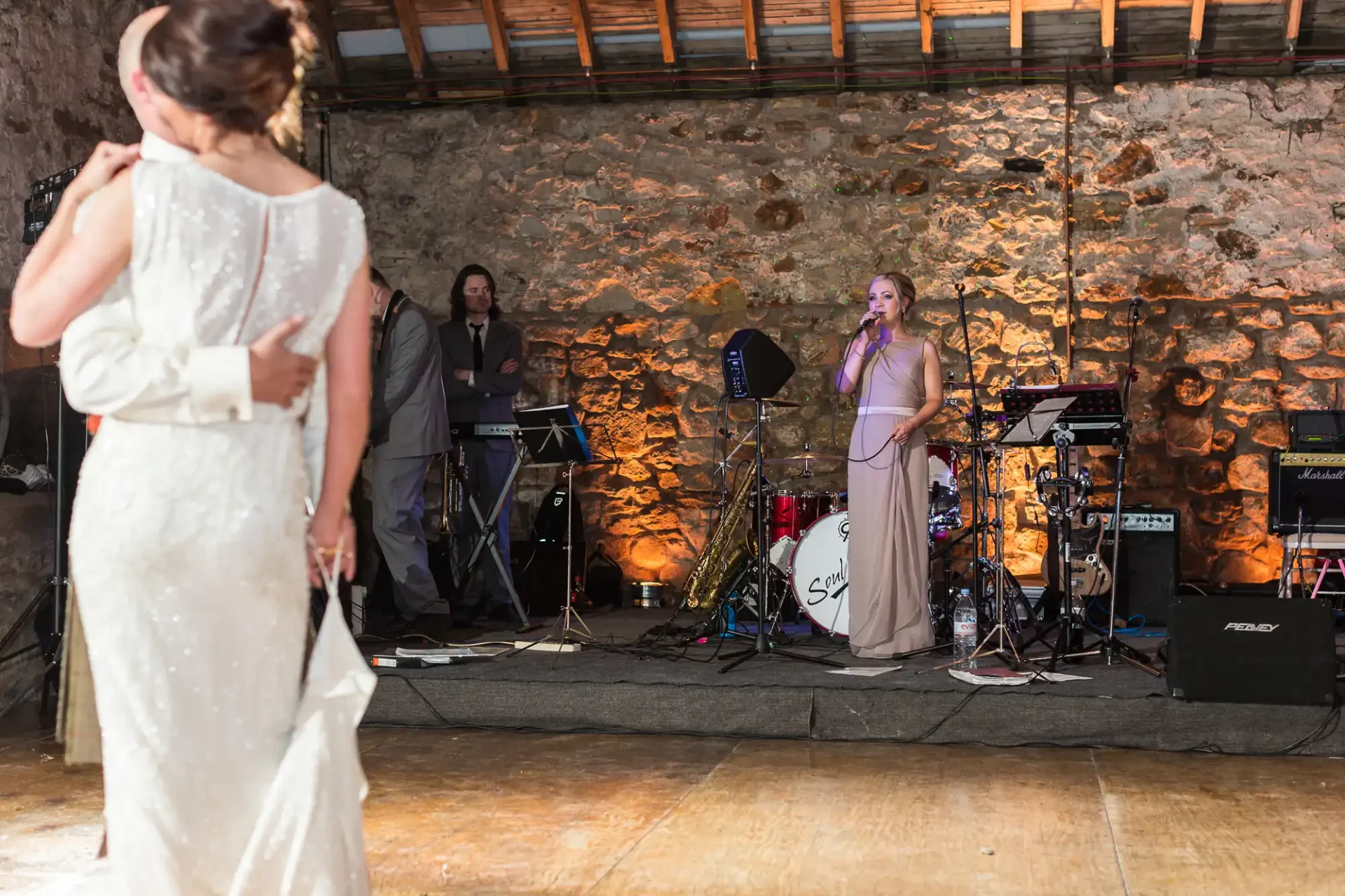 A bride listens to a female singer performing with a band in a rustic venue with stone walls.