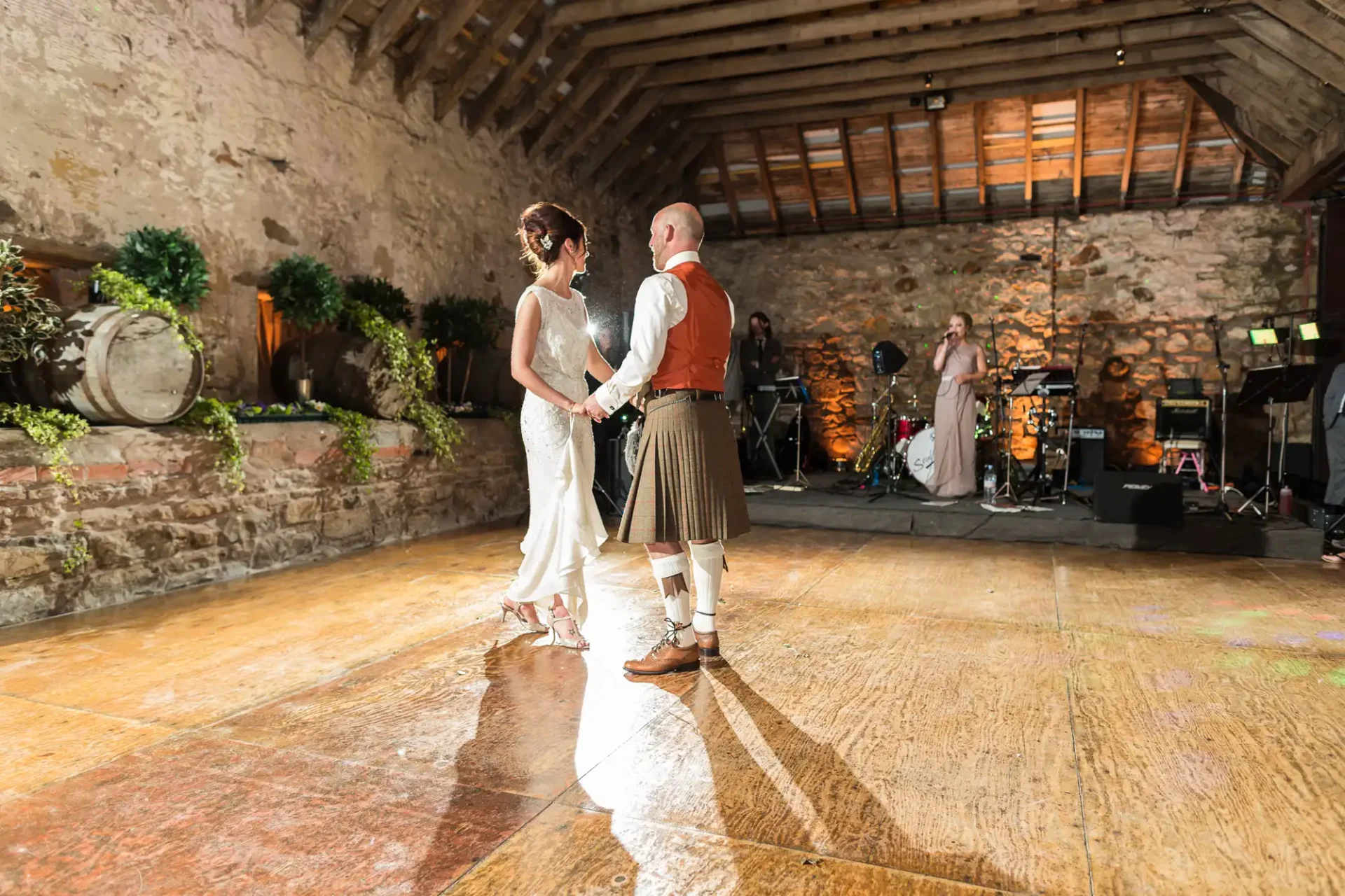 A bride and groom in traditional scottish attire share a dance in a rustic barn venue with a live band in the background.