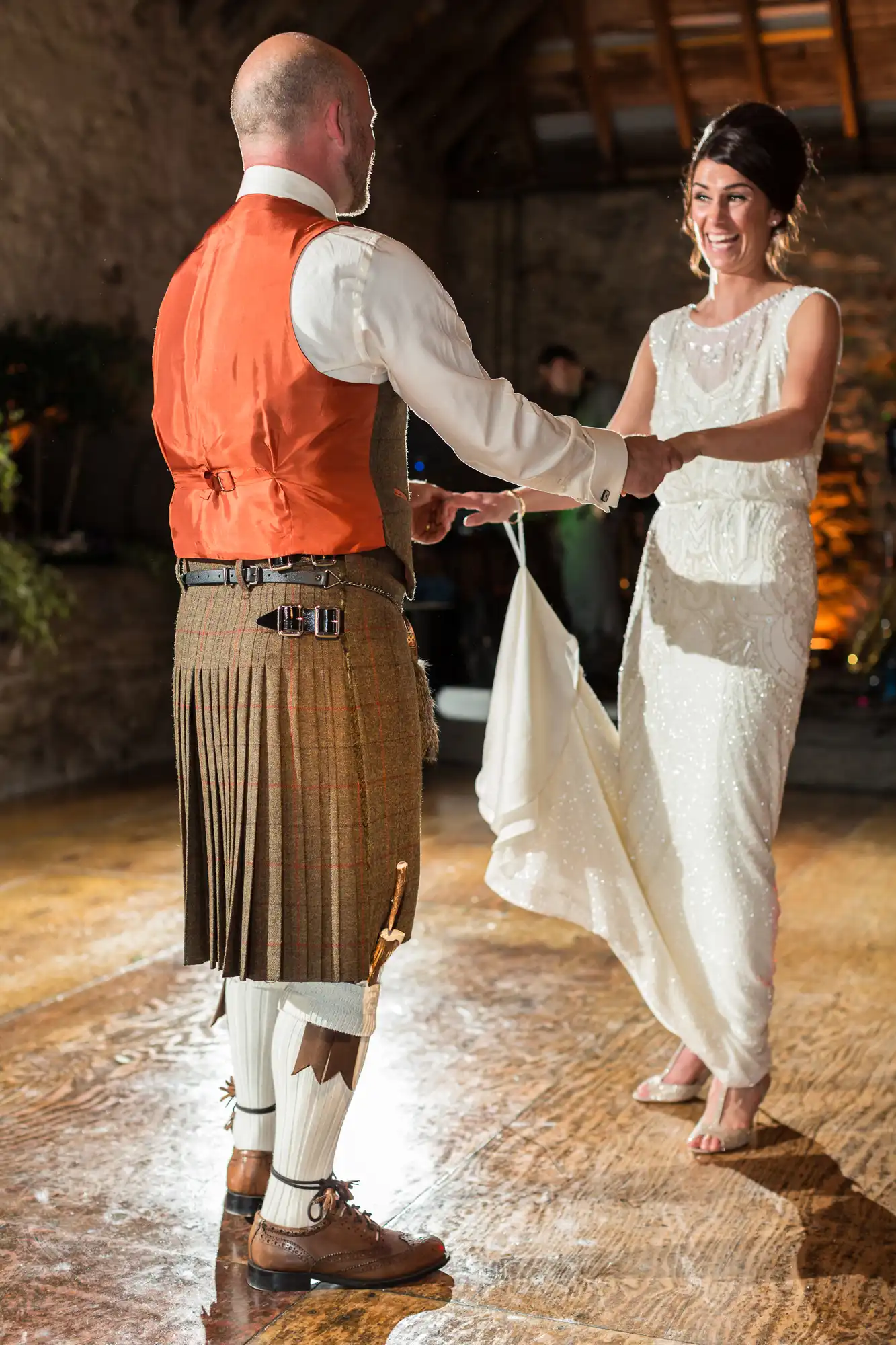 A bride and groom in traditional scottish attire dance joyfully at their wedding, the groom wearing a kilt and the bride in a white dress.