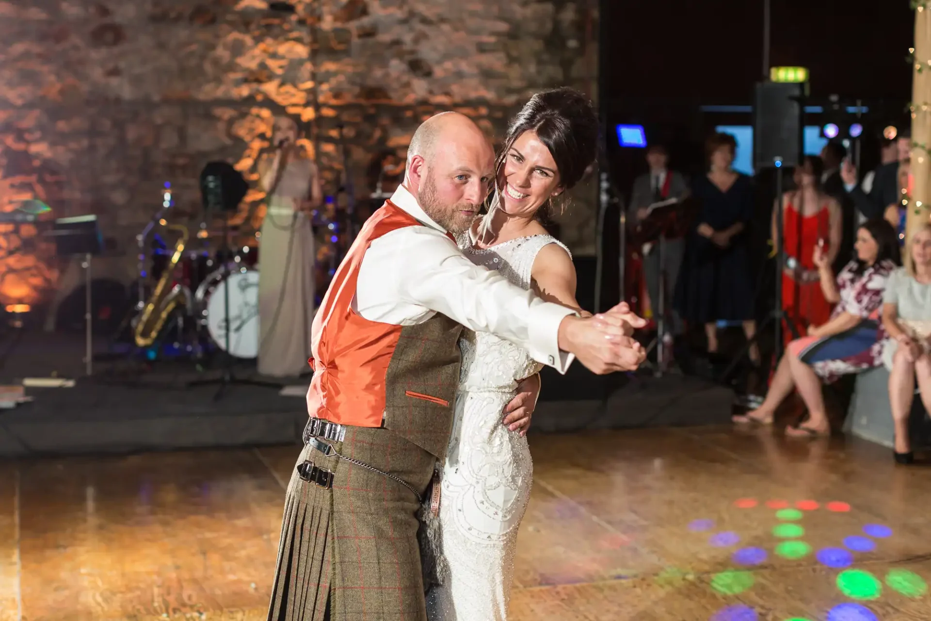 A couple dances closely at a wedding reception, the man in a kilt and the woman in a white dress, with guests and a band in the background.