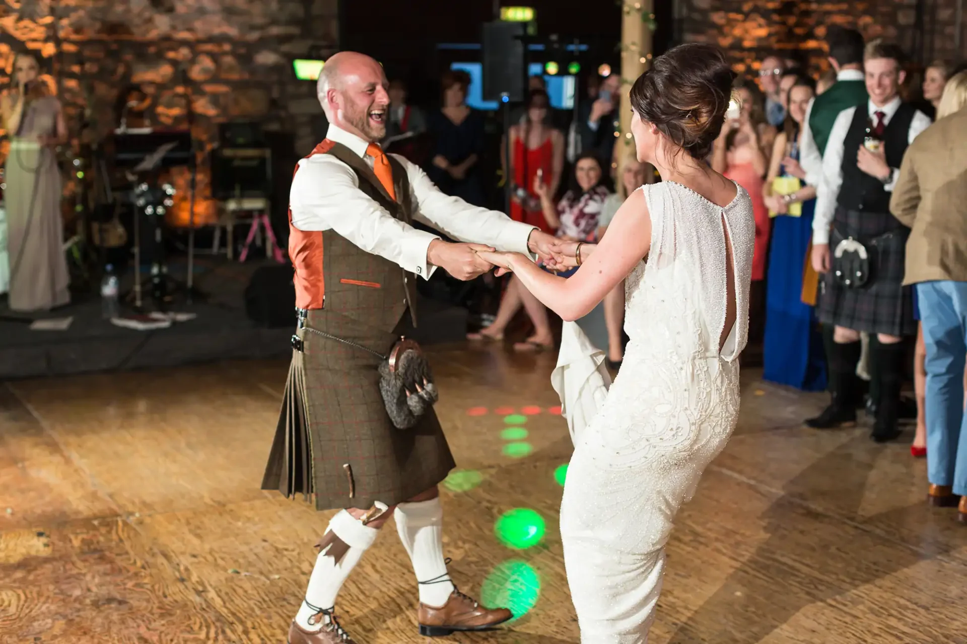 Bride and groom joyfully dancing in a reception hall, with the groom wearing a kilt and the guests watching.