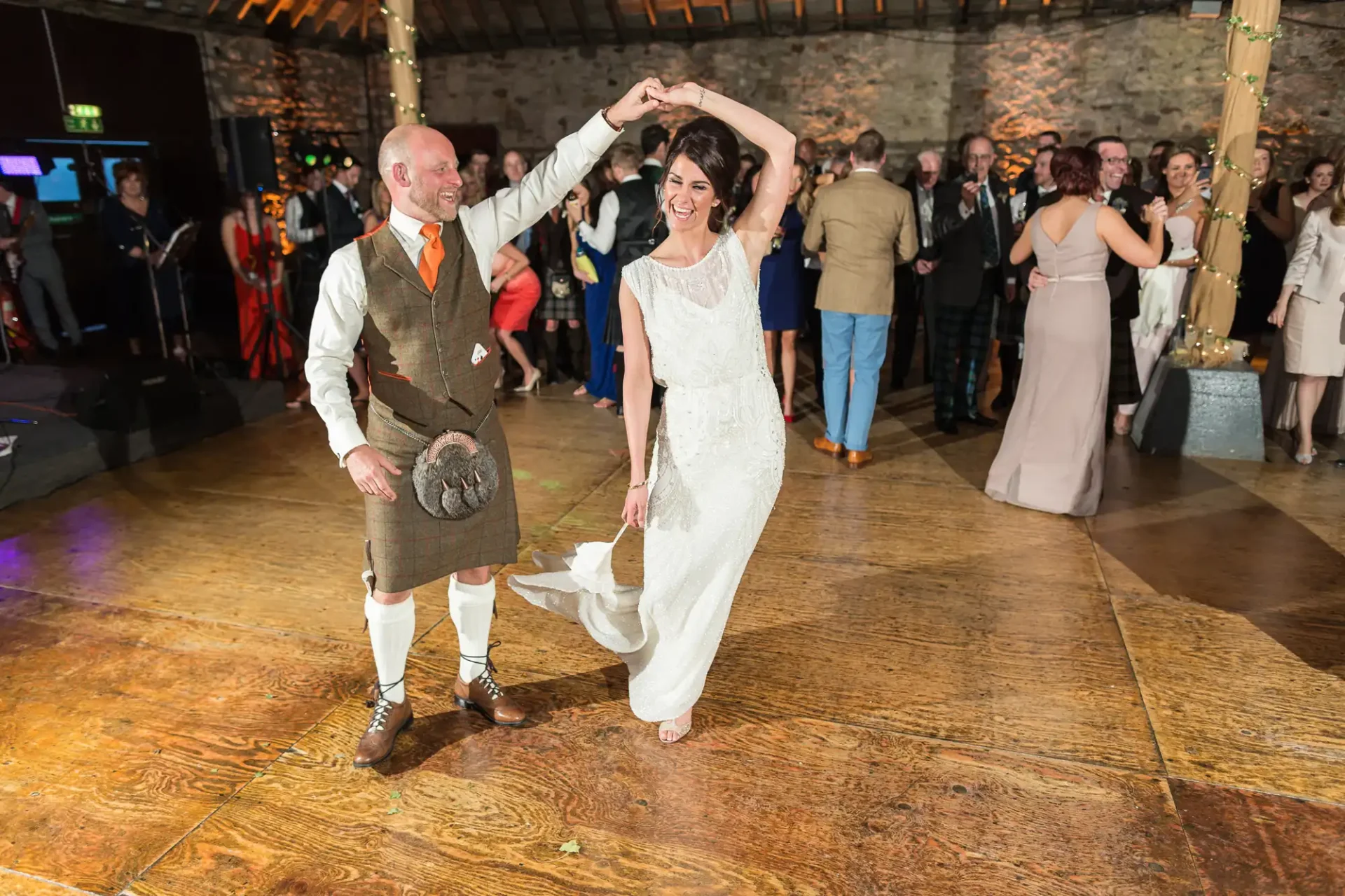 A bride and groom in traditional scottish attire dancing joyfully at their wedding reception, surrounded by guests.