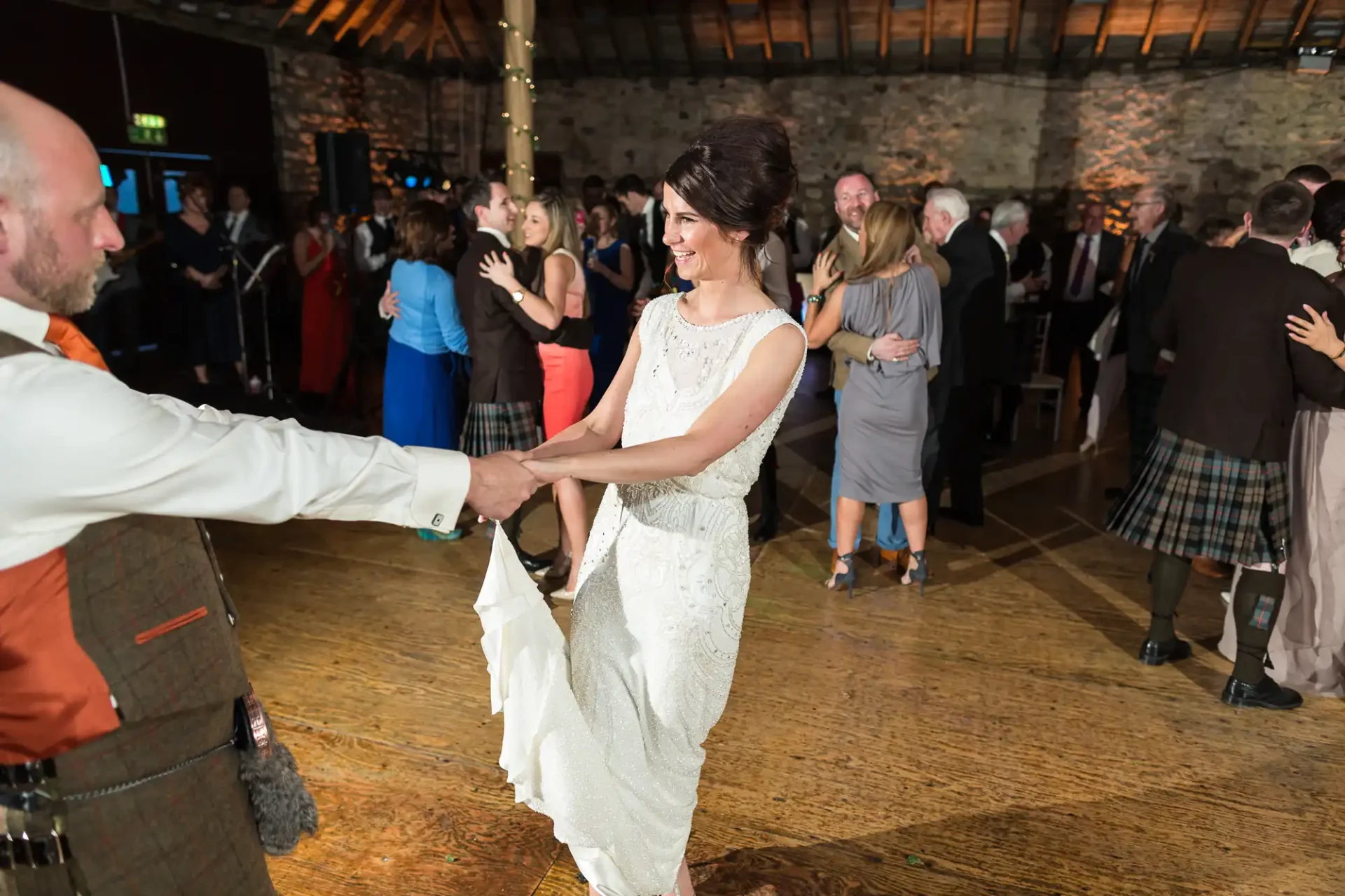 A woman in a white dress dances joyfully with a man in traditional scottish attire at a lively indoor event.