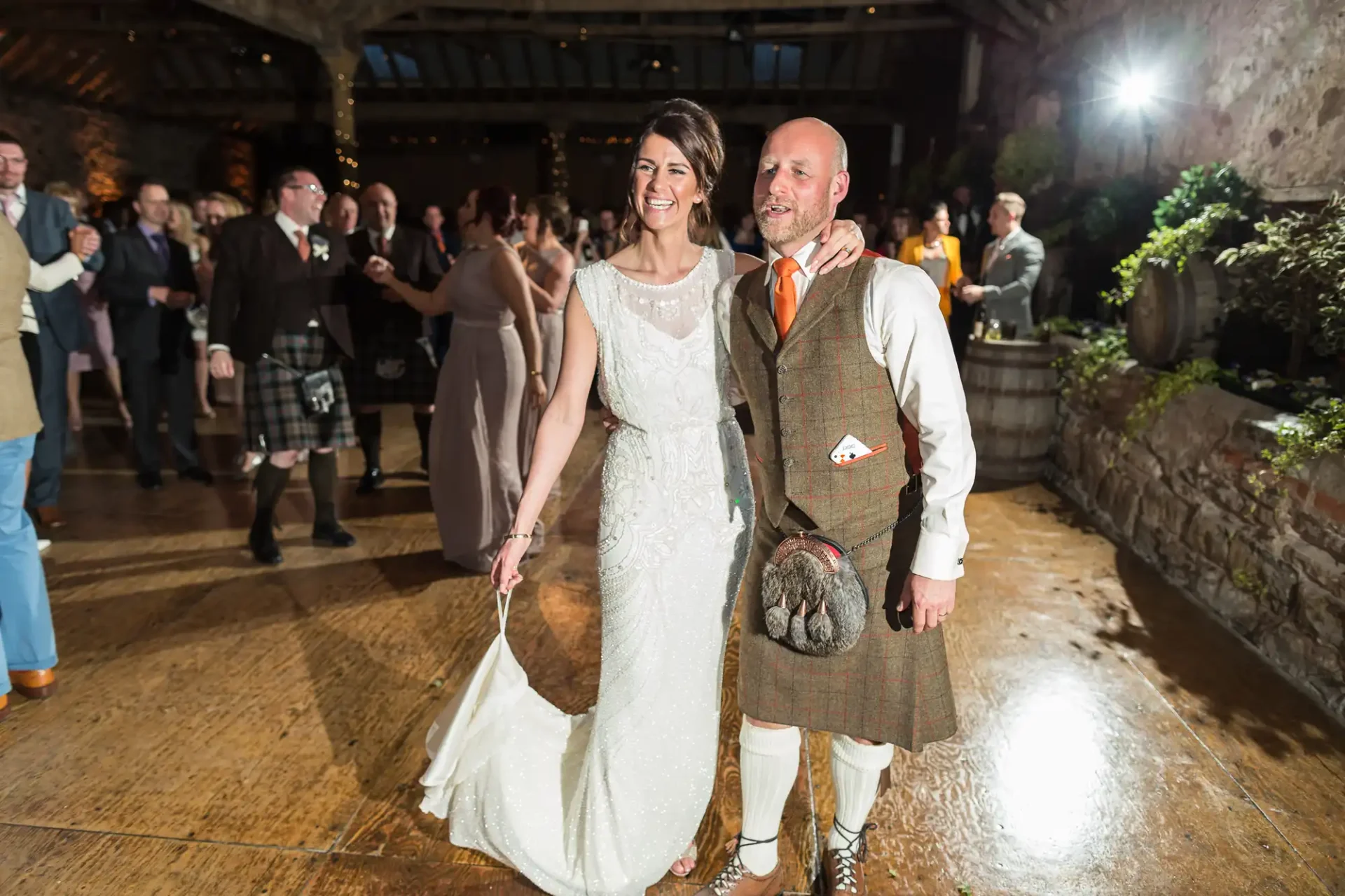 A bride in a white lace dress and a groom in a kilt and tweed jacket walk hand in hand, smiling at a wedding reception.