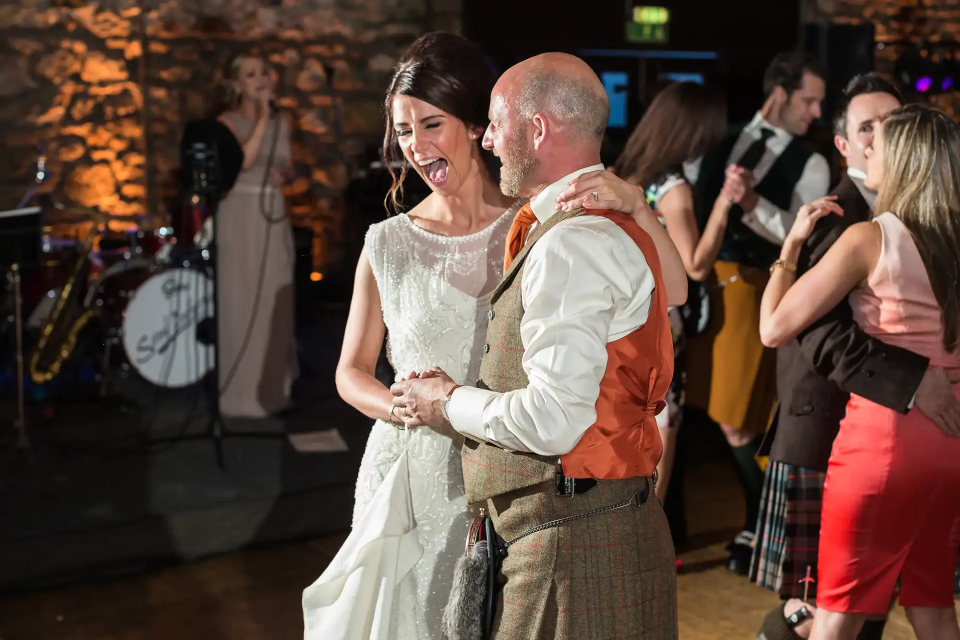A bride and groom in wedding attire laugh together while dancing at a reception, with guests and a band in the background.