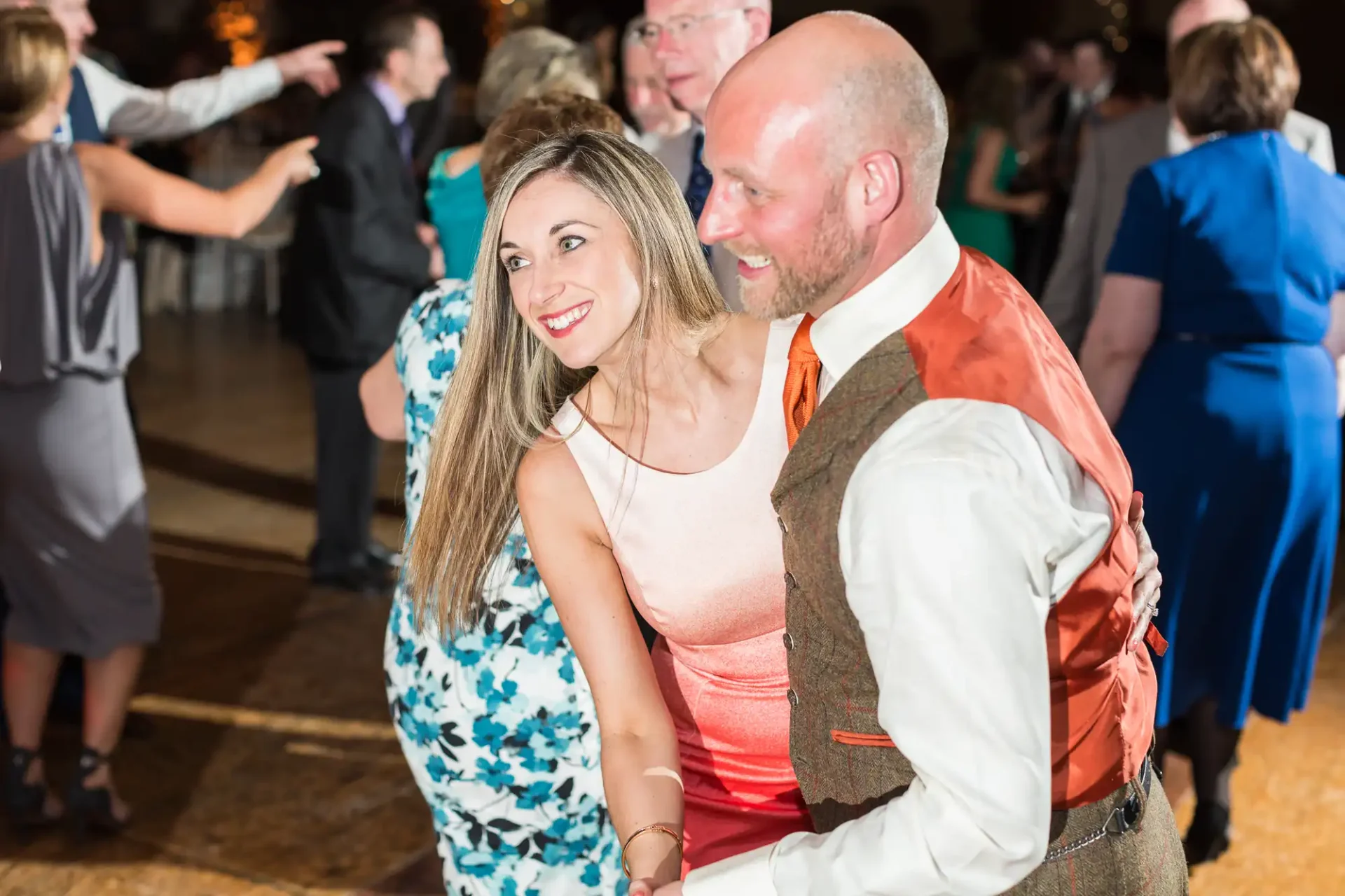A couple smiles while dancing at a lively wedding reception, surrounded by other guests. the man wears a vest and the woman a sleeveless dress.