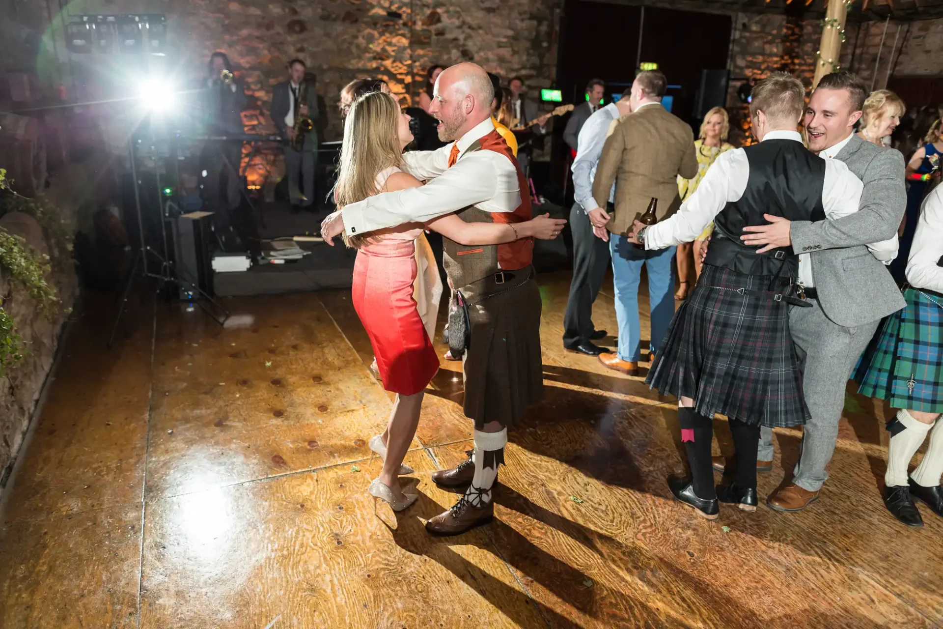 Couples dancing in a lively setting, one man wearing traditional scottish attire, with a band playing in the background.