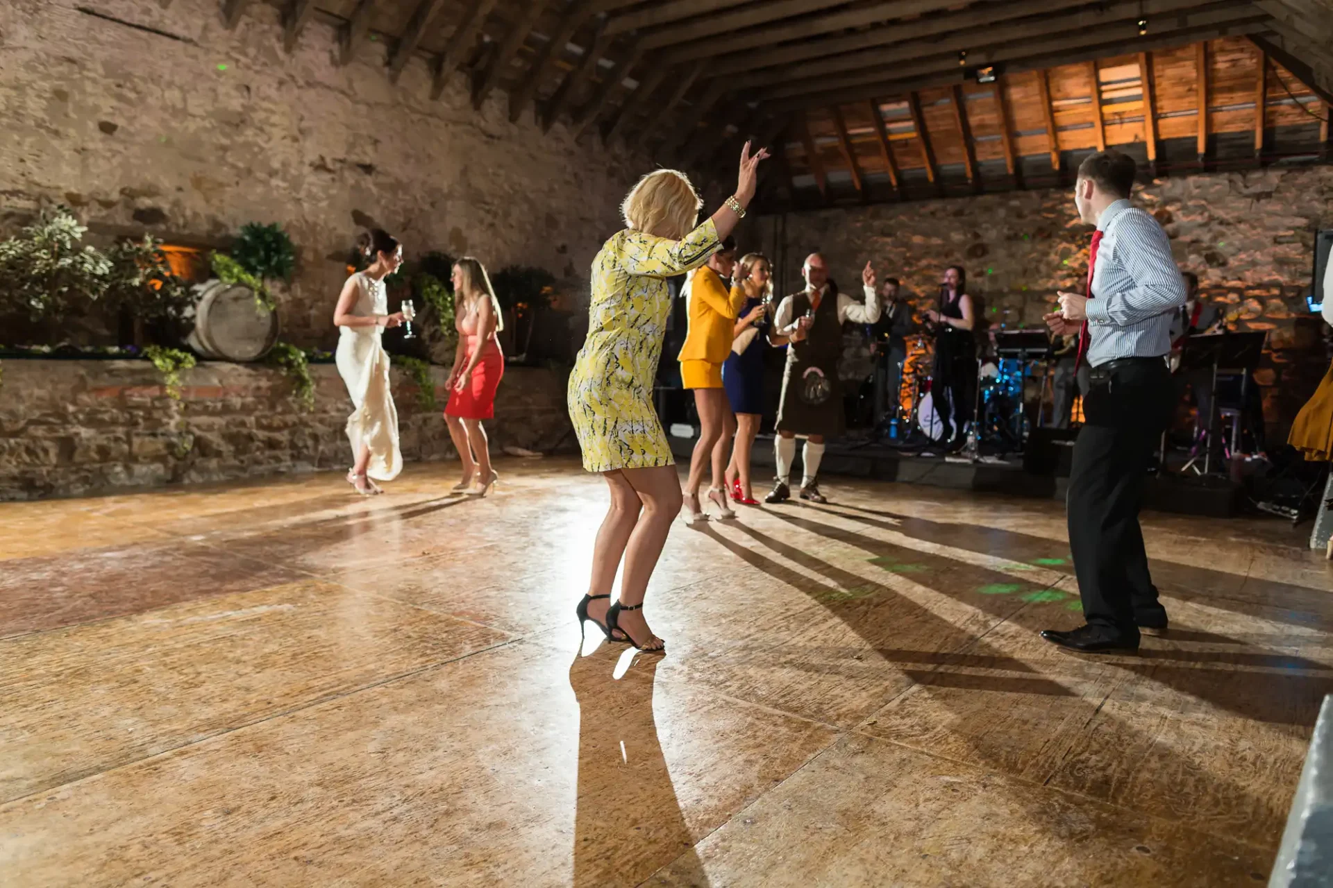 Guests dancing enthusiastically on a wooden floor at an indoor party, with a live band playing in the background.