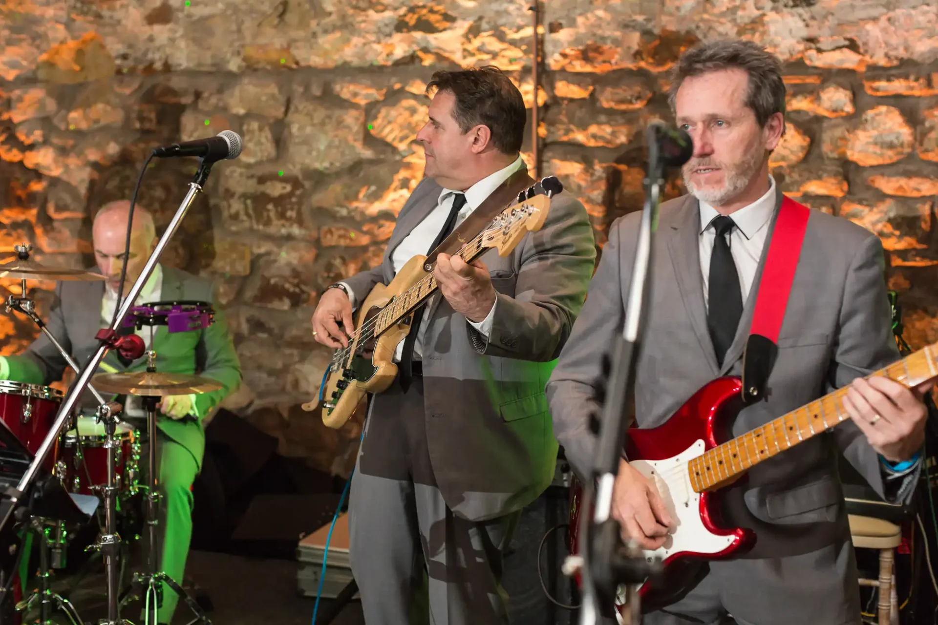 Three middle-aged men perform in a band at an indoor event, playing drums and electric guitars, against a rustic stone wall backdrop.