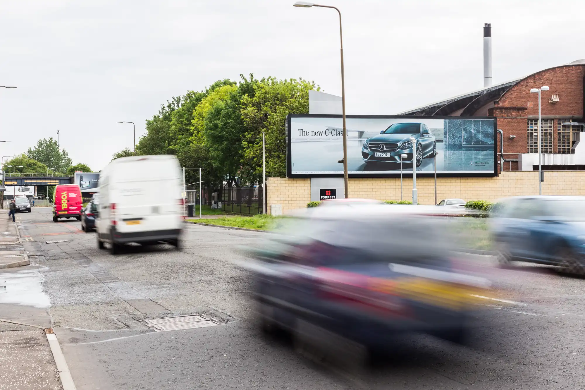 A busy road with several moving vehicles, a white van, and other cars. A large billboard advertises a new car model. Trees and buildings are visible in the background.