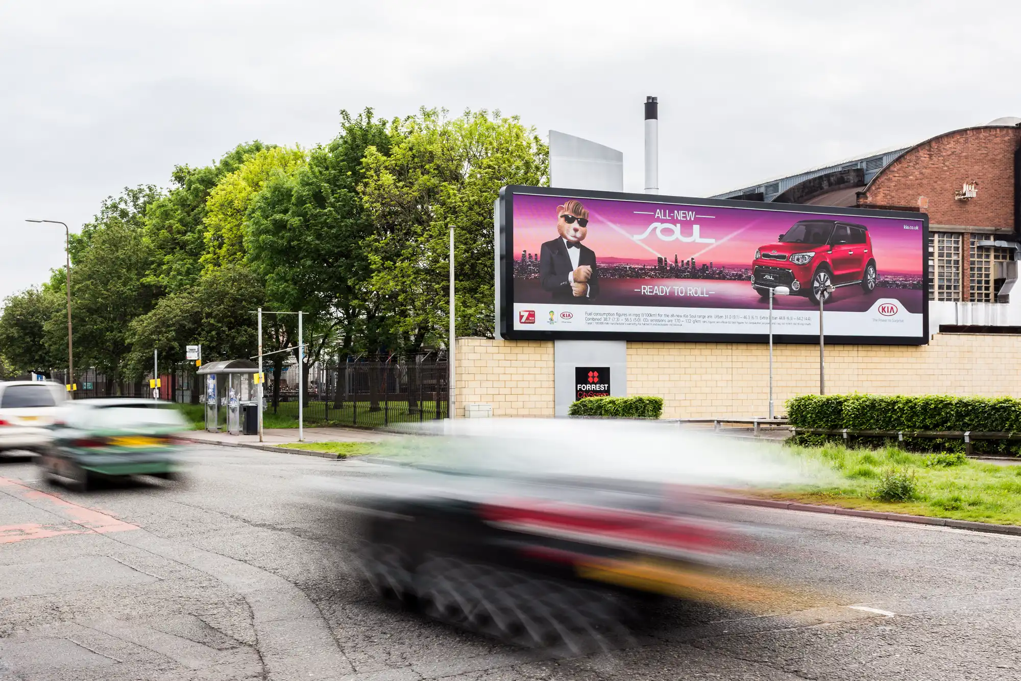 Street with a billboard advertisement for the "All-New Soul" car model. Vehicles are blurred due to motion. The scene includes greenery and some buildings in the background.