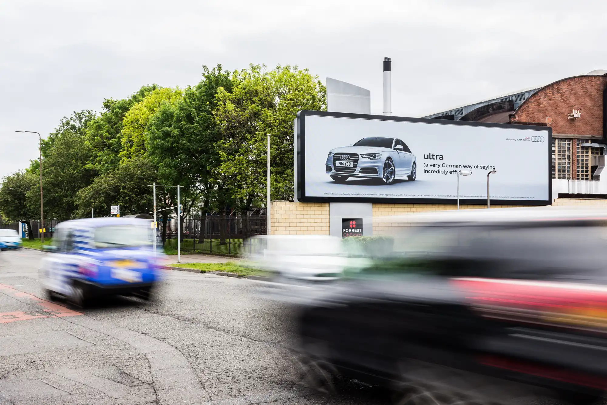 A billboard with an Audi advertisement depicting a silver car and the text "ultra" is displayed beside a road with several blurry, moving vehicles. Trees and a building are in the background.