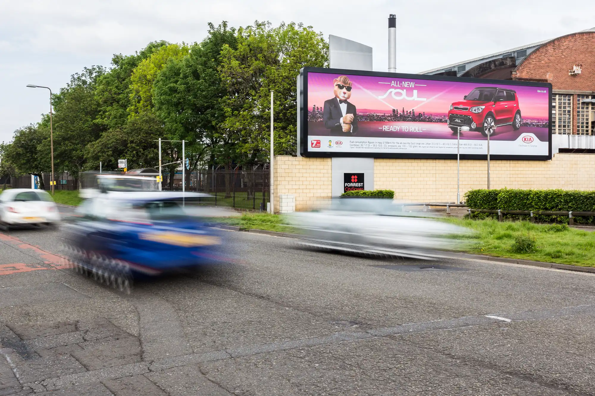 Vehicles in motion on a road with a large billboard in the background advertising the "All-New Soul" car model. Trees and buildings are visible behind the billboard.