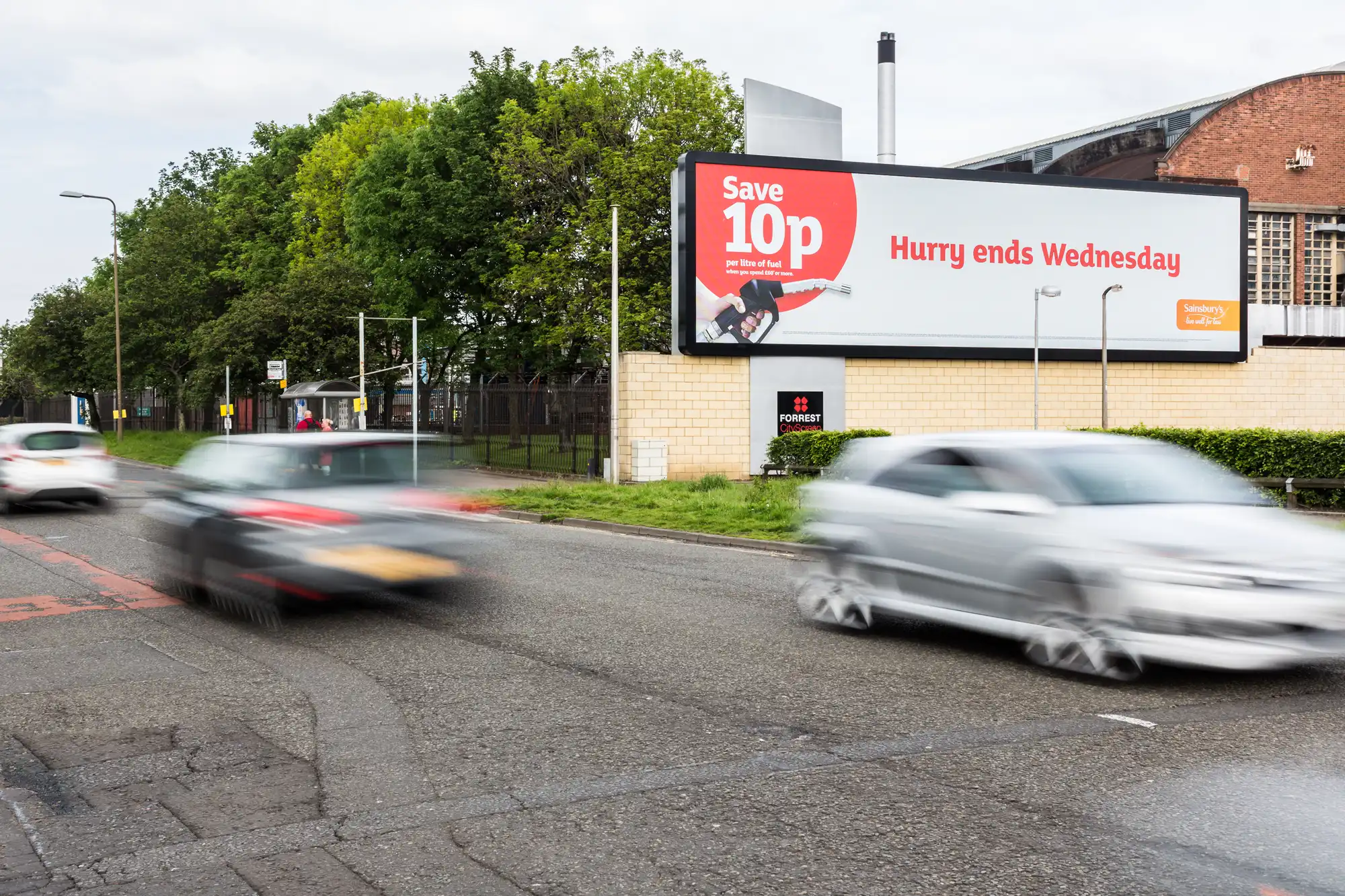 Traffic passes by a billboard advertising a 10p discount that ends on Wednesday, with blurred vehicles in the foreground and trees in the background.