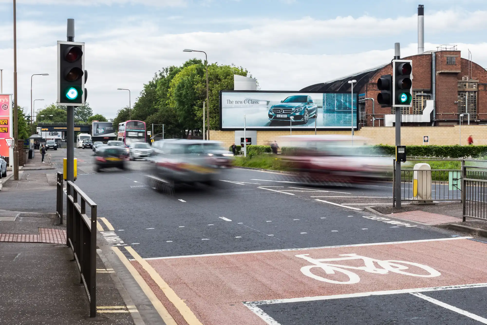 A busy intersection with cars in motion under a green traffic light, a bicycle lane in the foreground, and a billboard advertising a car in the background.