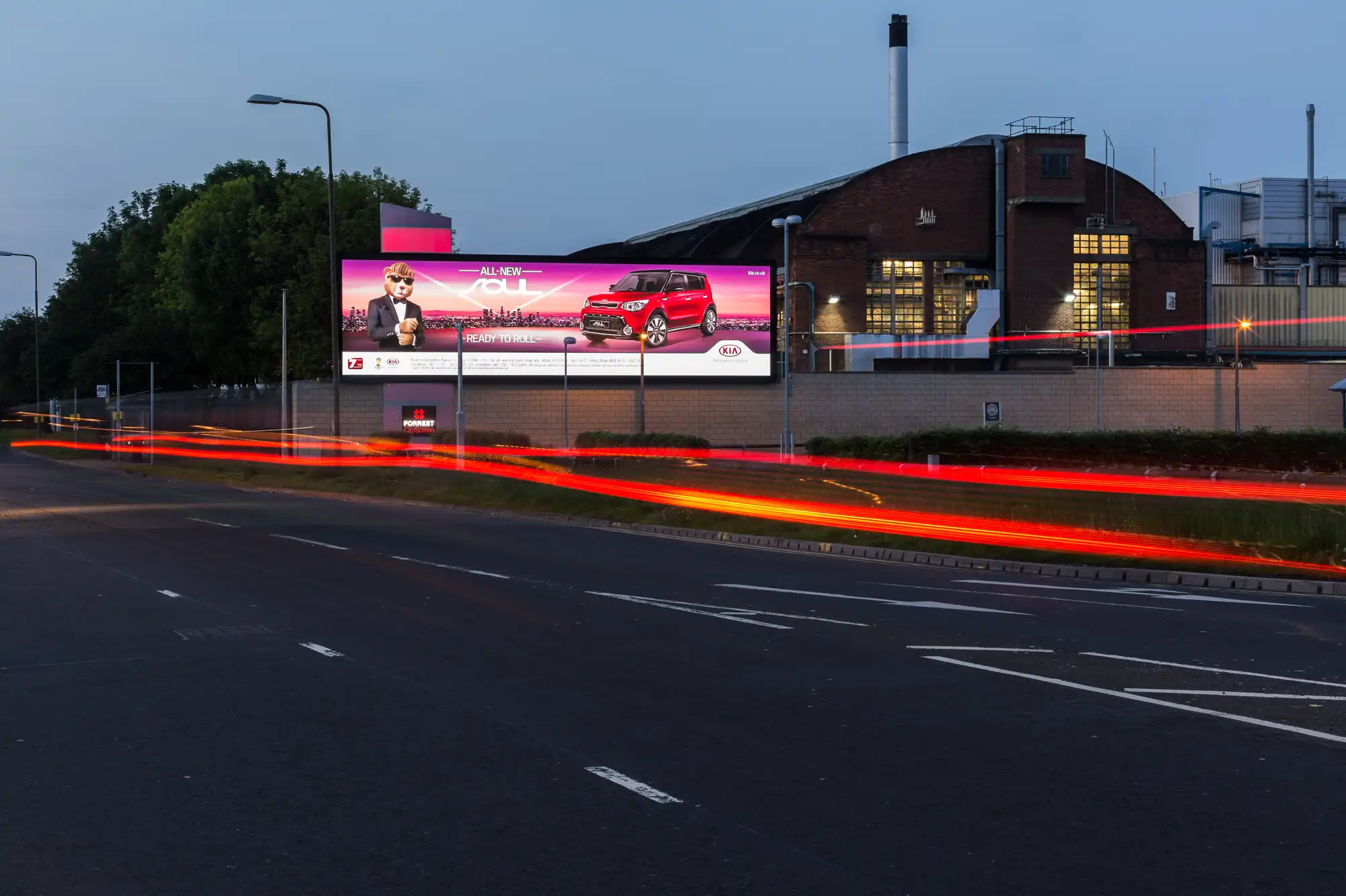 Billboard advertisement for a car on a roadside with visible light trails from passing vehicles in the foreground. An industrial building is in the background during twilight.