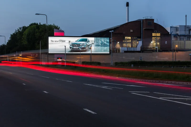 Nighttime view of a road with light trails from passing vehicles; a large billboard on a building displays an advertisement for a new car model.