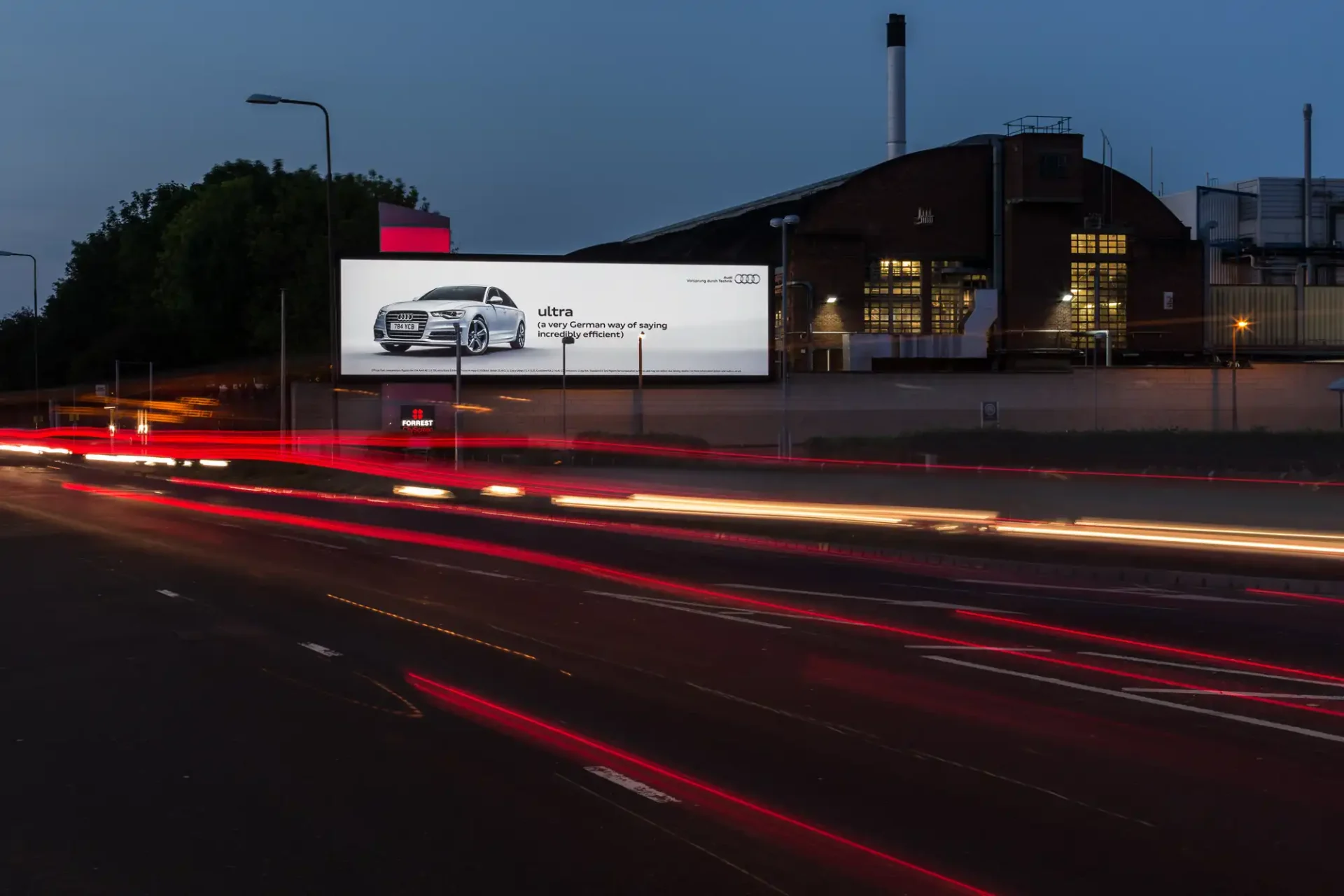 Product photographer in Edinburgh - D3 LED City Gateway Stevenson Road: A long exposure photo of a street with red tail lights from moving vehicles and a large illuminated billboard advertising an Audi car at dusk. An industrial building is in the background.