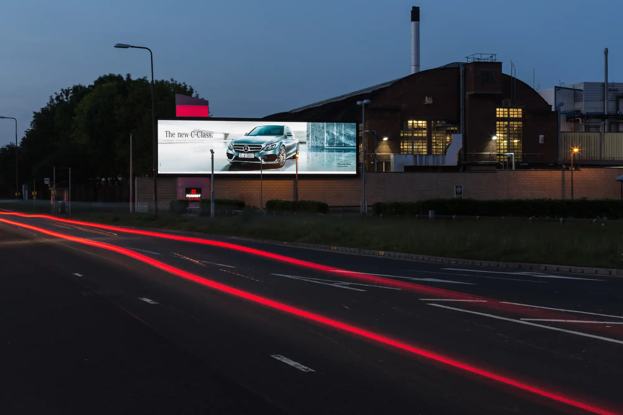 A billboard with a car advertisement is illuminated at dusk along a road with light trails from passing vehicles. An industrial building is seen in the background.