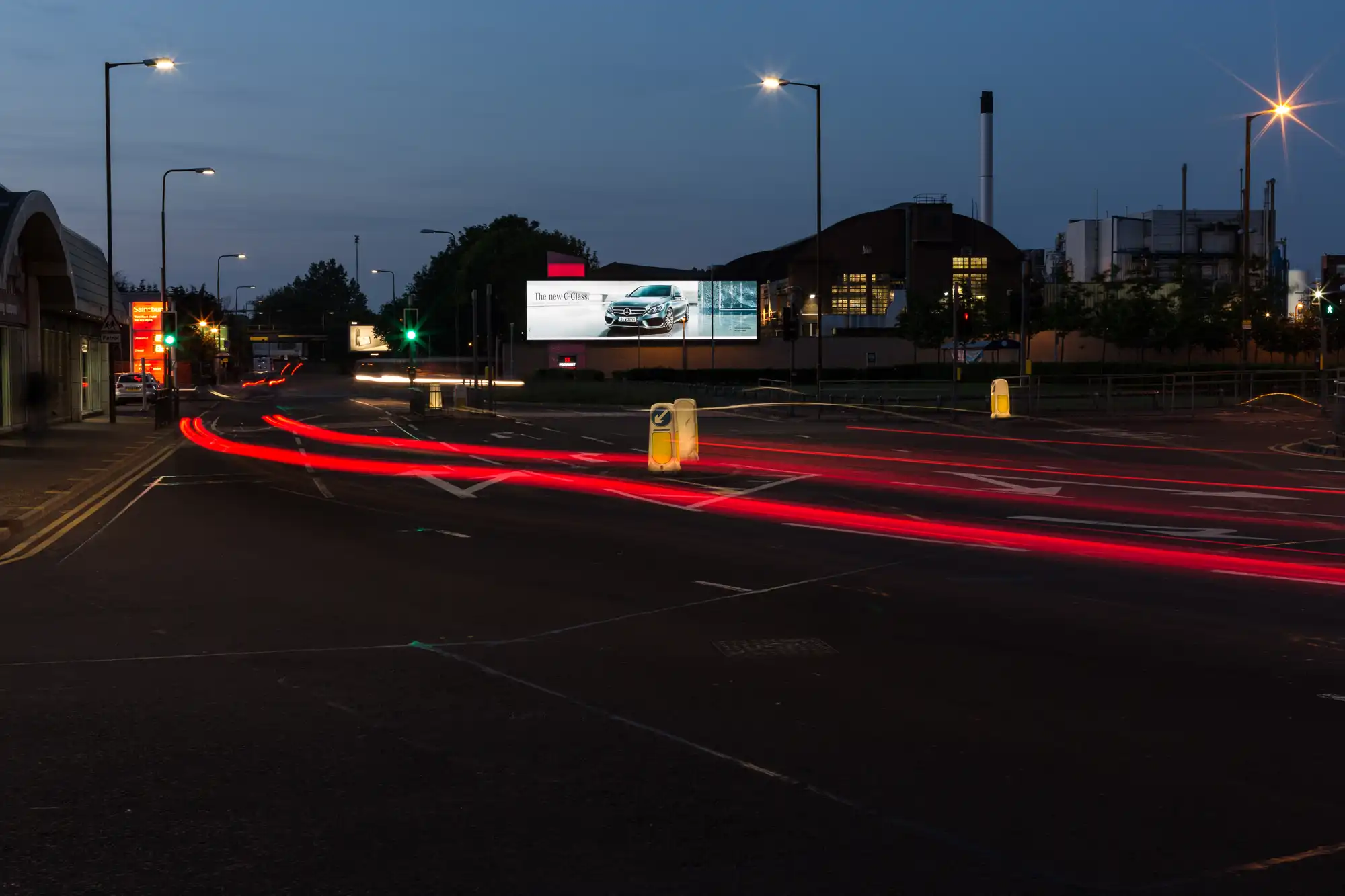 Nighttime view of a road with light trails from passing vehicles and a lit billboard in the background.