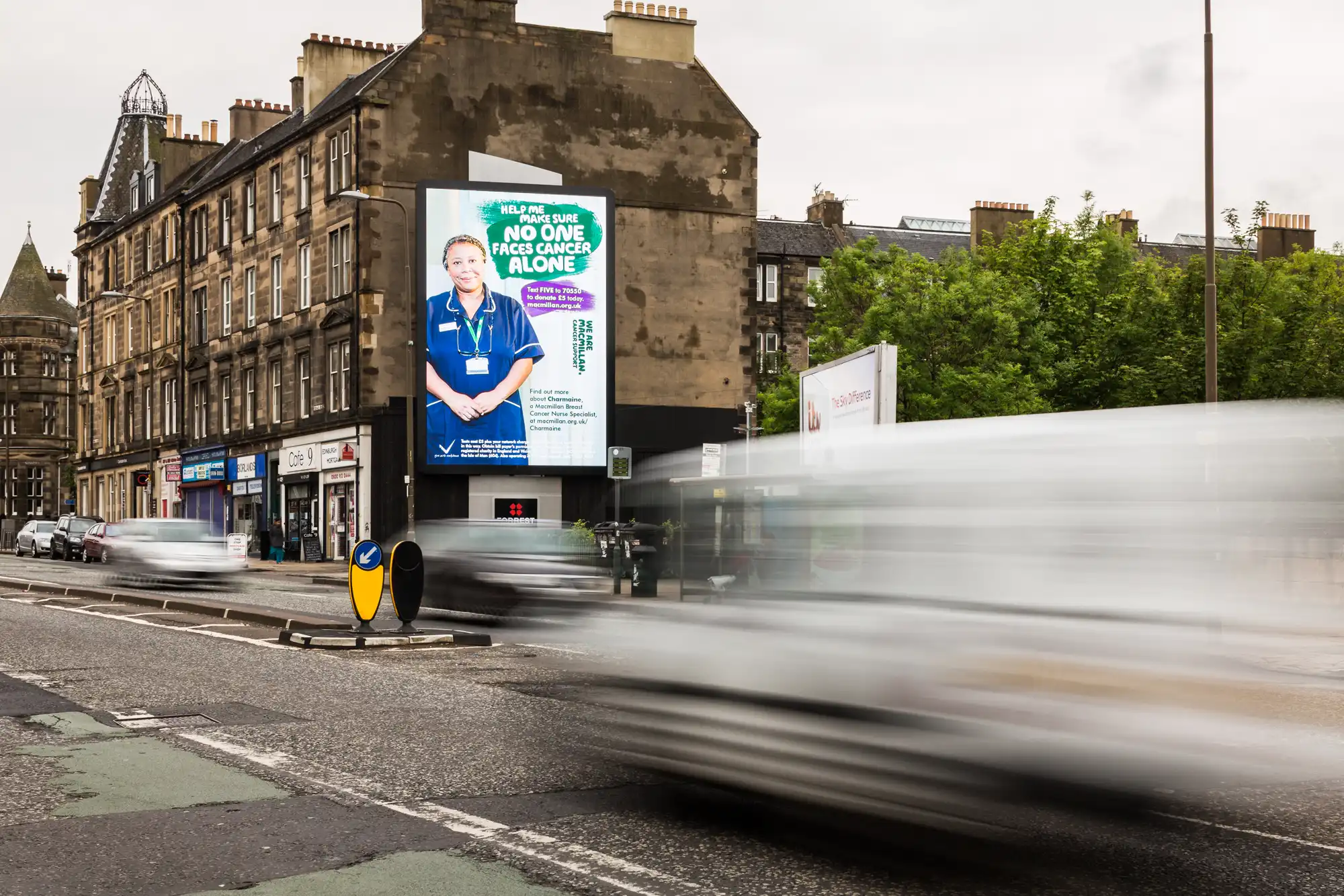 Busy street with blurred traffic, large billboard on building displays healthcare message "No one faces cancer alone" featuring a smiling nurse. Brick buildings and greenery in the background.