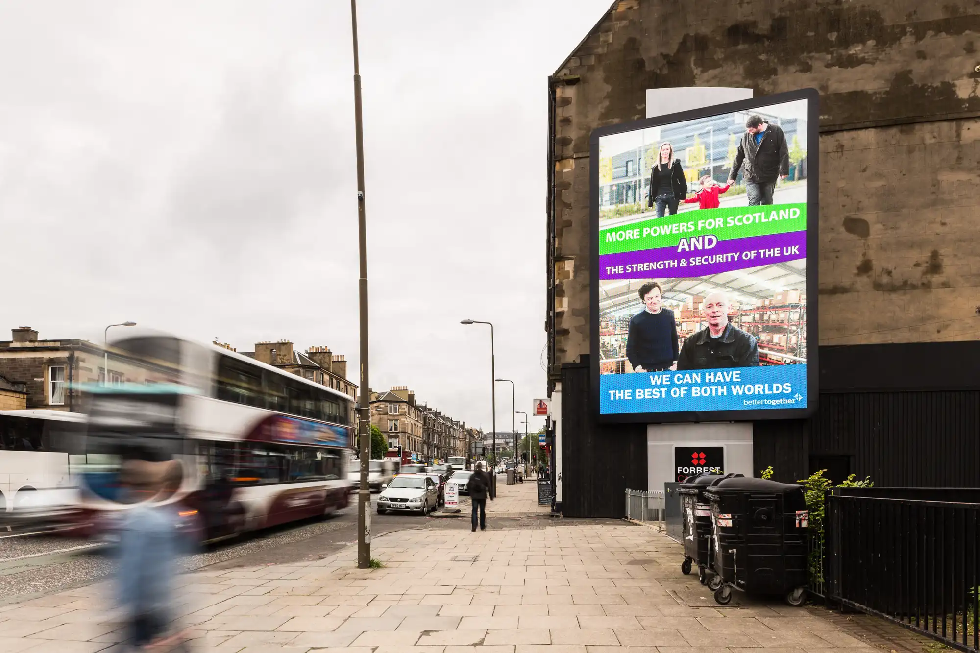 A street view showing a large political billboard. The billboard features two images of people and text promoting more powers for Scotland and the strength and security of the UK. A bus drives by.