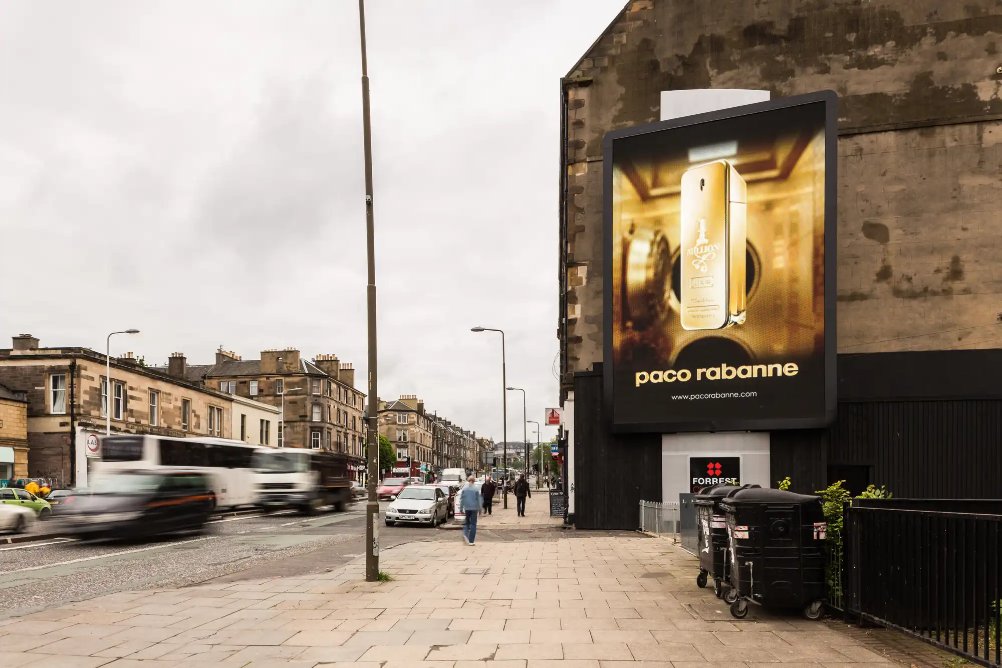 Street view of a large billboard advertising Paco Rabanne's "1 Million" fragrance on a busy urban sidewalk with pedestrians and a passing car.