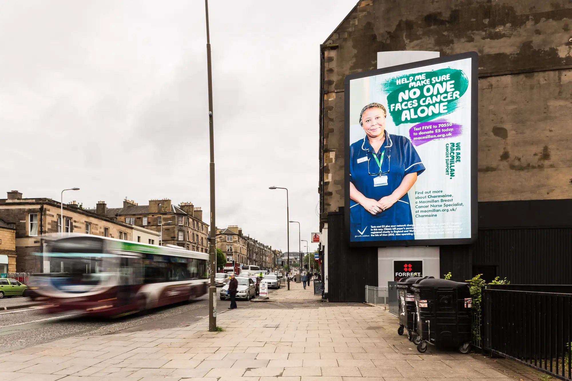 A digital billboard on a street displays an advertisement featuring a medical professional promoting cancer support services. A bus and moving traffic are visible on the left, with buildings in the background.