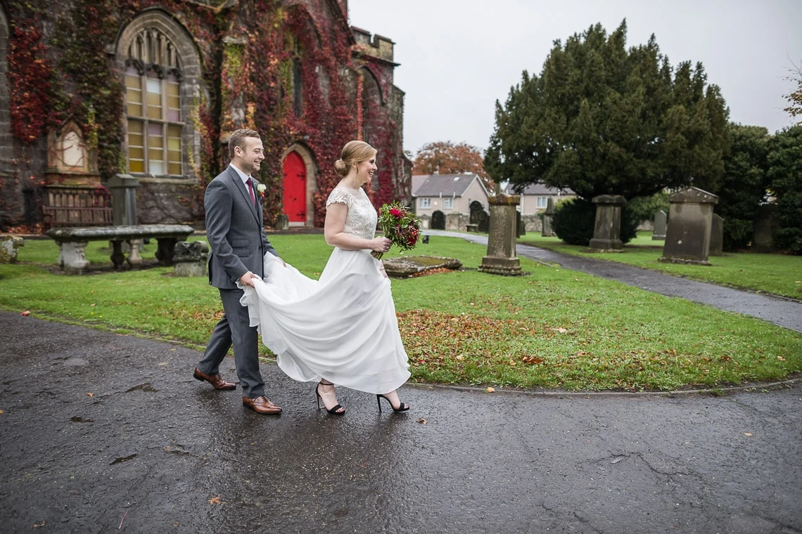 groom helps his bride walk through the grounds of Liberton Kirk