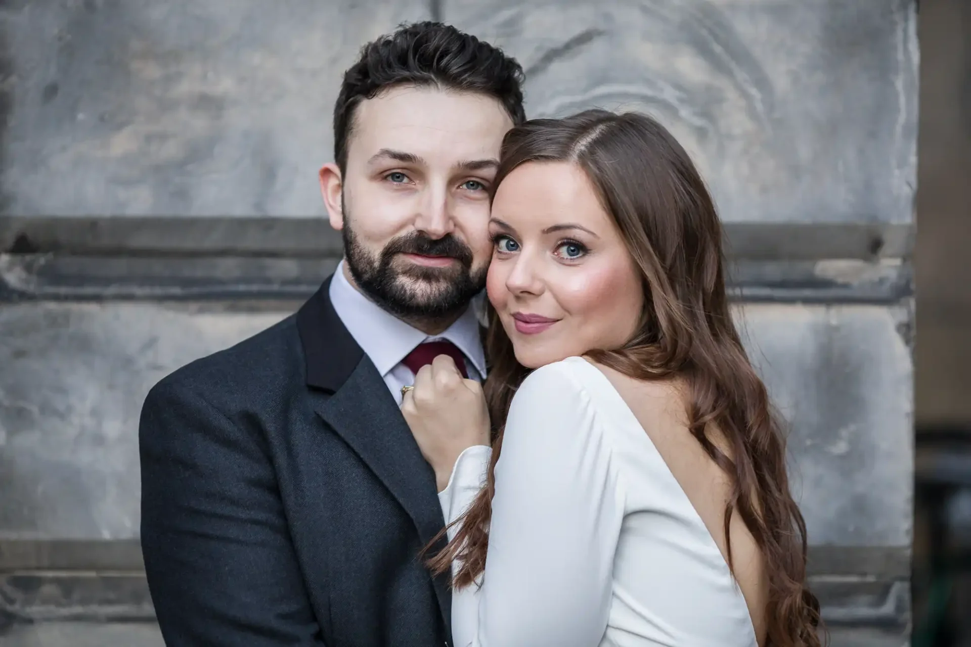 A couple poses closely, with the woman in a white dress and the man in a dark suit, against a stone wall backdrop.