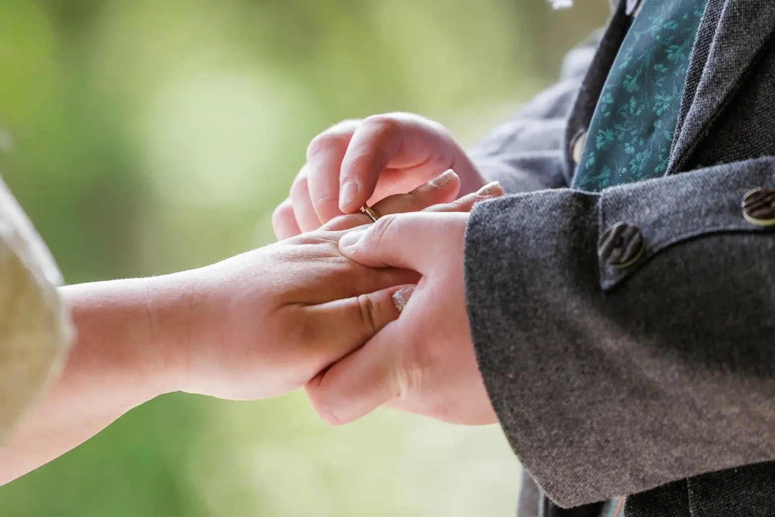 Close-up of one person placing a ring on another person's finger, both wearing formal attire, with a blurred green background.