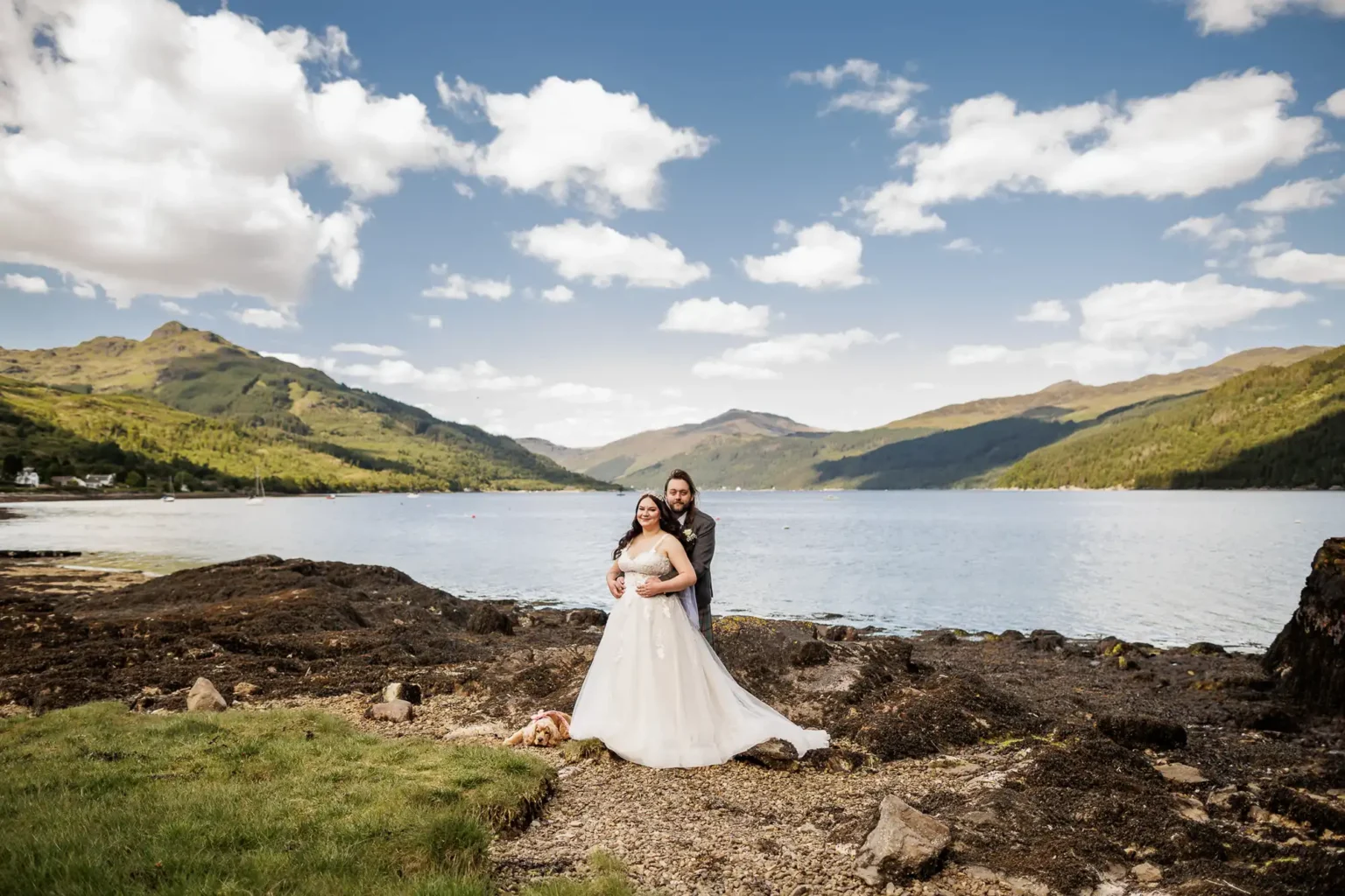 A bride and groom stand together by a lake with mountains in the background under a partly cloudy sky.