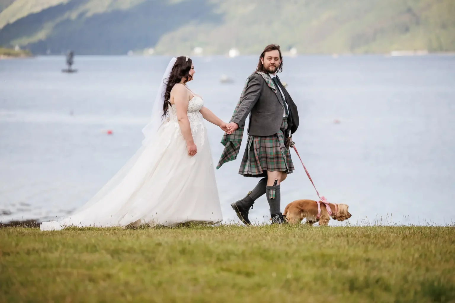 A bride and groom in wedding attire, with the groom in a kilt, walk hand-in-hand by a lakeside, accompanied by a small dog on a leash.