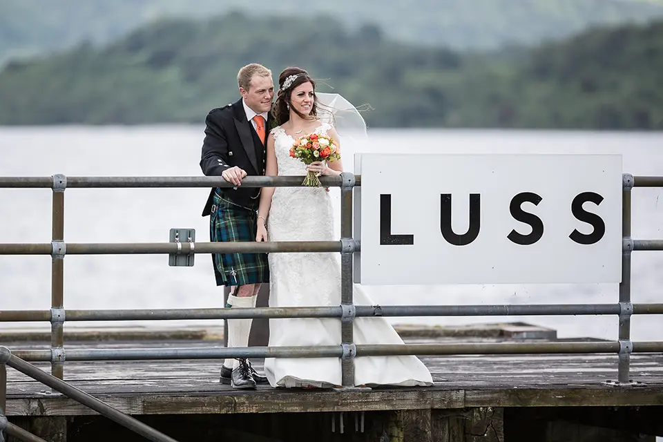 A bride and groom stand together on a wooden pier in luss, overlooking a lake, with the groom wearing a kilt and the bride in a white gown, holding a bouquet.