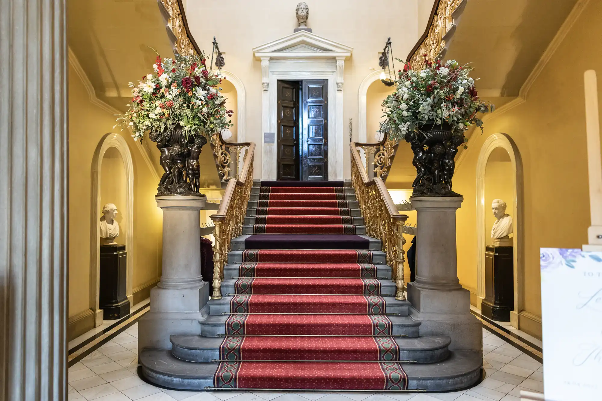A grand staircase with red carpet, ornate banisters, and floral arrangements on each side. Bust sculptures are displayed in niches along the walls, under high ceilings with elegant decor.
