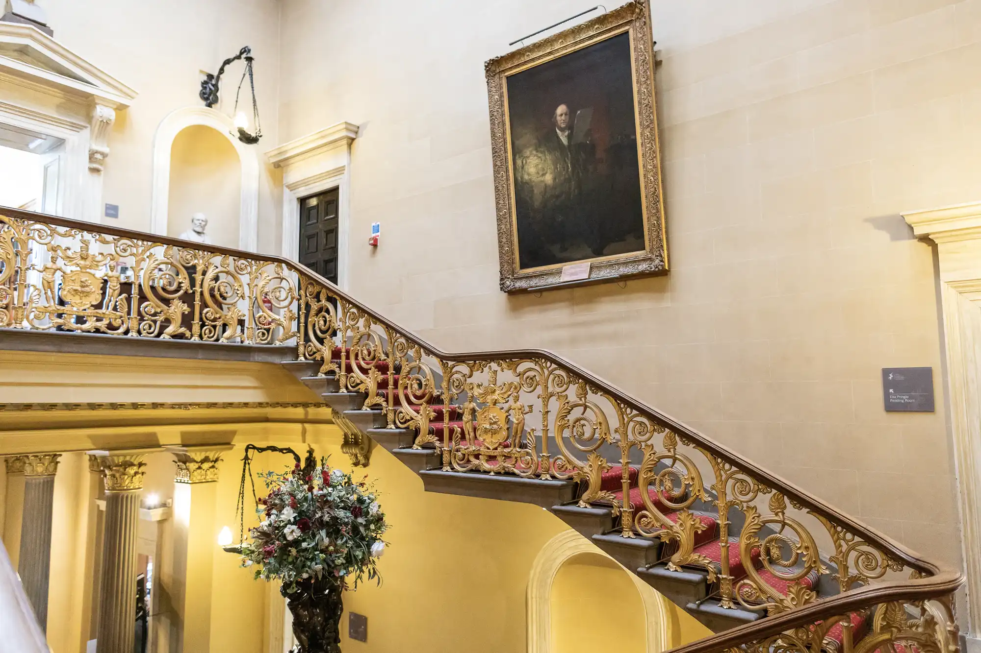 Photo of an ornate staircase with a gold railing and red carpeting in an old building. A large portrait hangs on the wall above the staircase, and a decorative floral arrangement sits by the stairs.